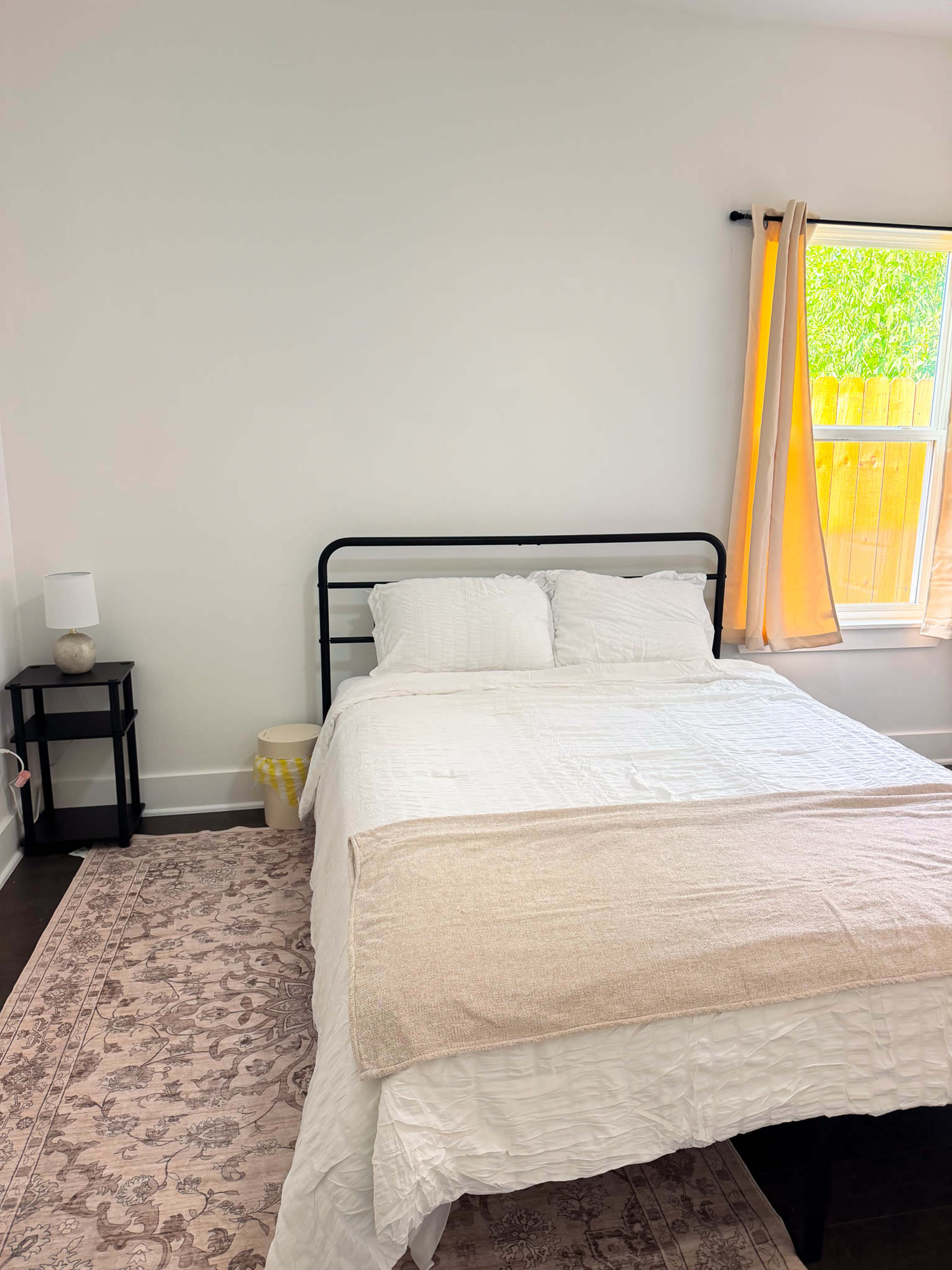 The image shows a neatly made bed with white bedding, positioned in a simple bedroom featuring a nightstand and a window with light curtains.