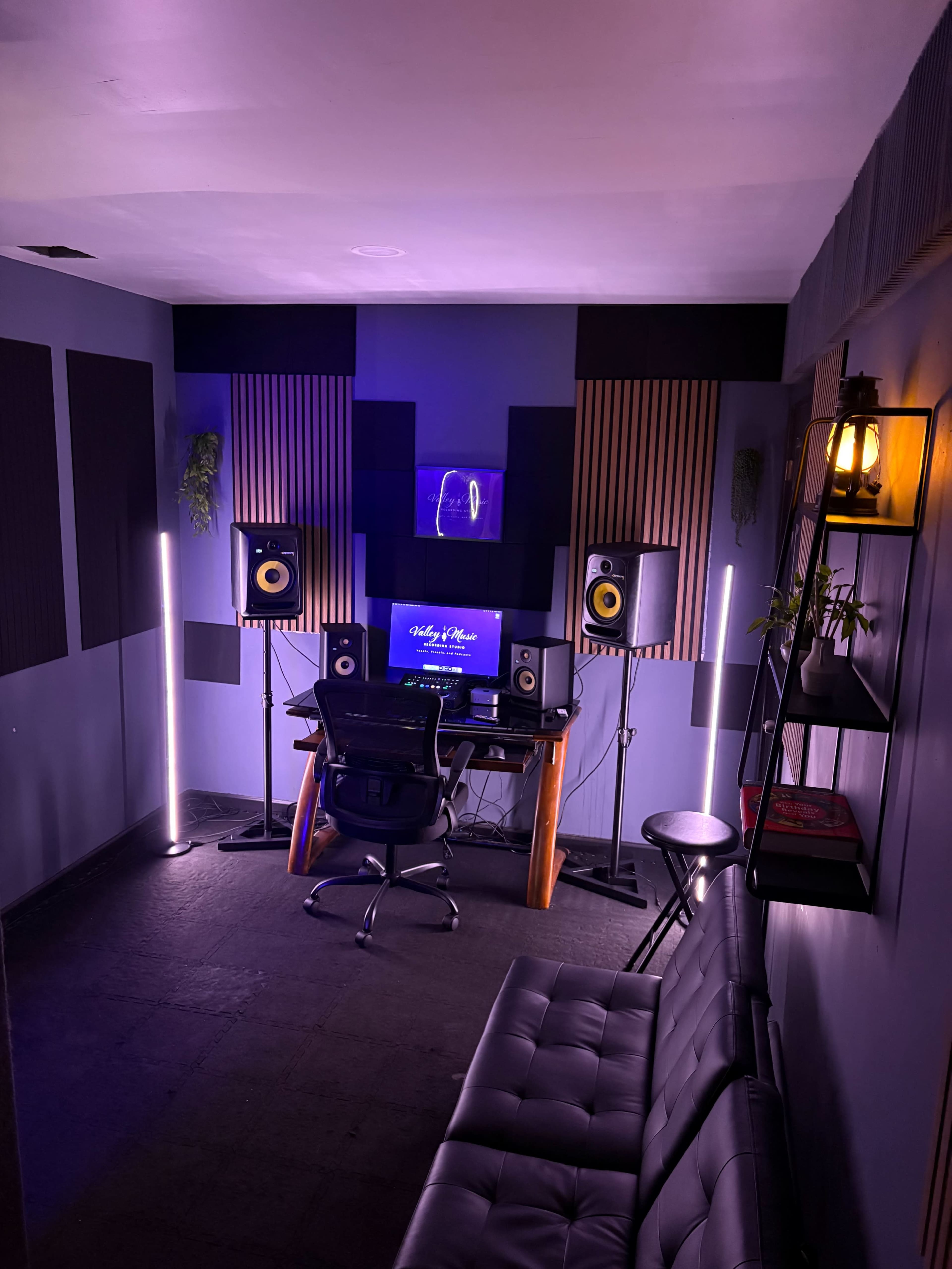 The image shows a modern music production studio featuring a desk with a computer and monitors, surrounded by acoustic panels and illuminated by soft lighting.