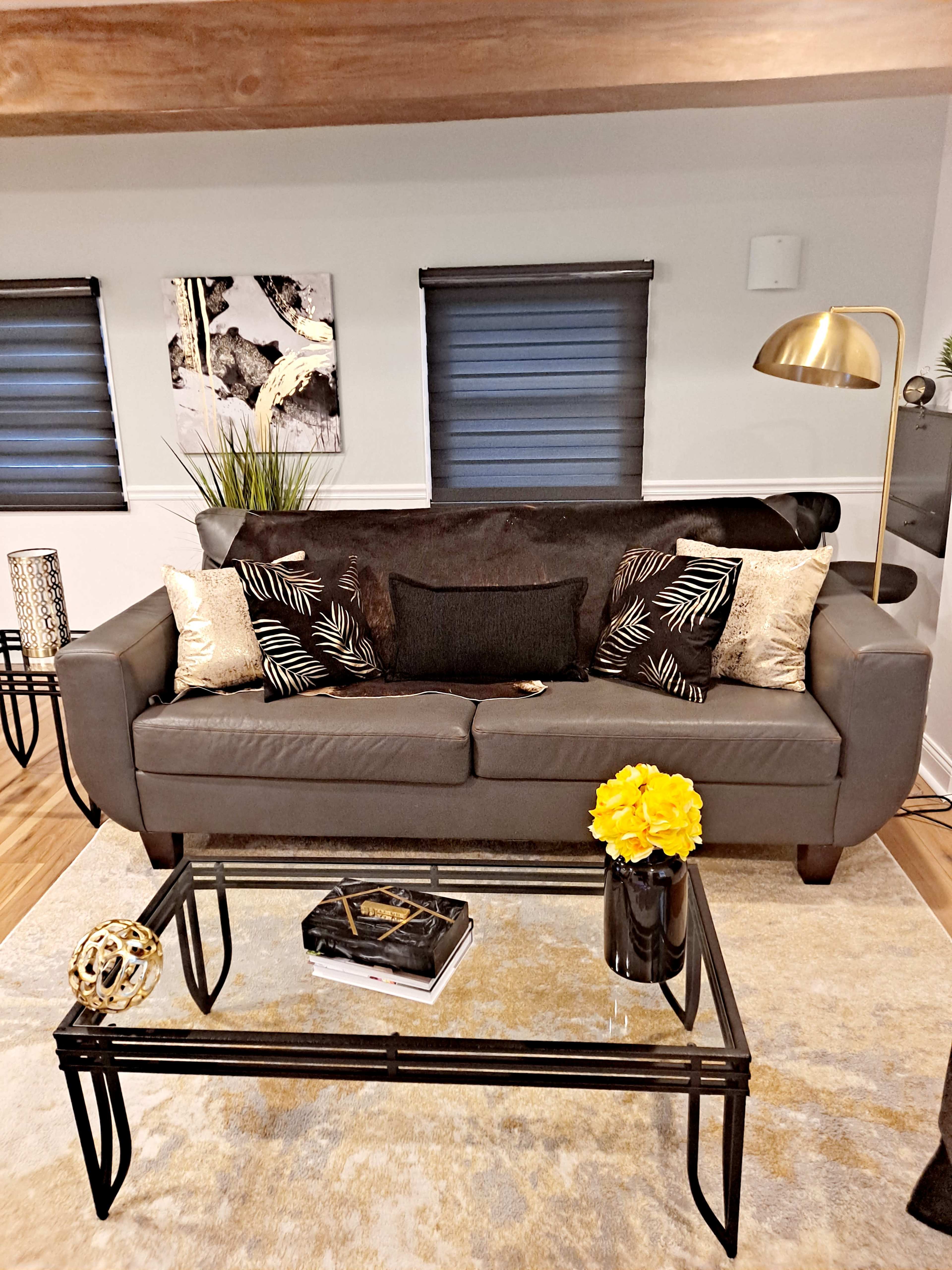 A gray sofa with decorative pillows sits in a living room next to a glass coffee table adorned with a book and a vase of yellow flowers.