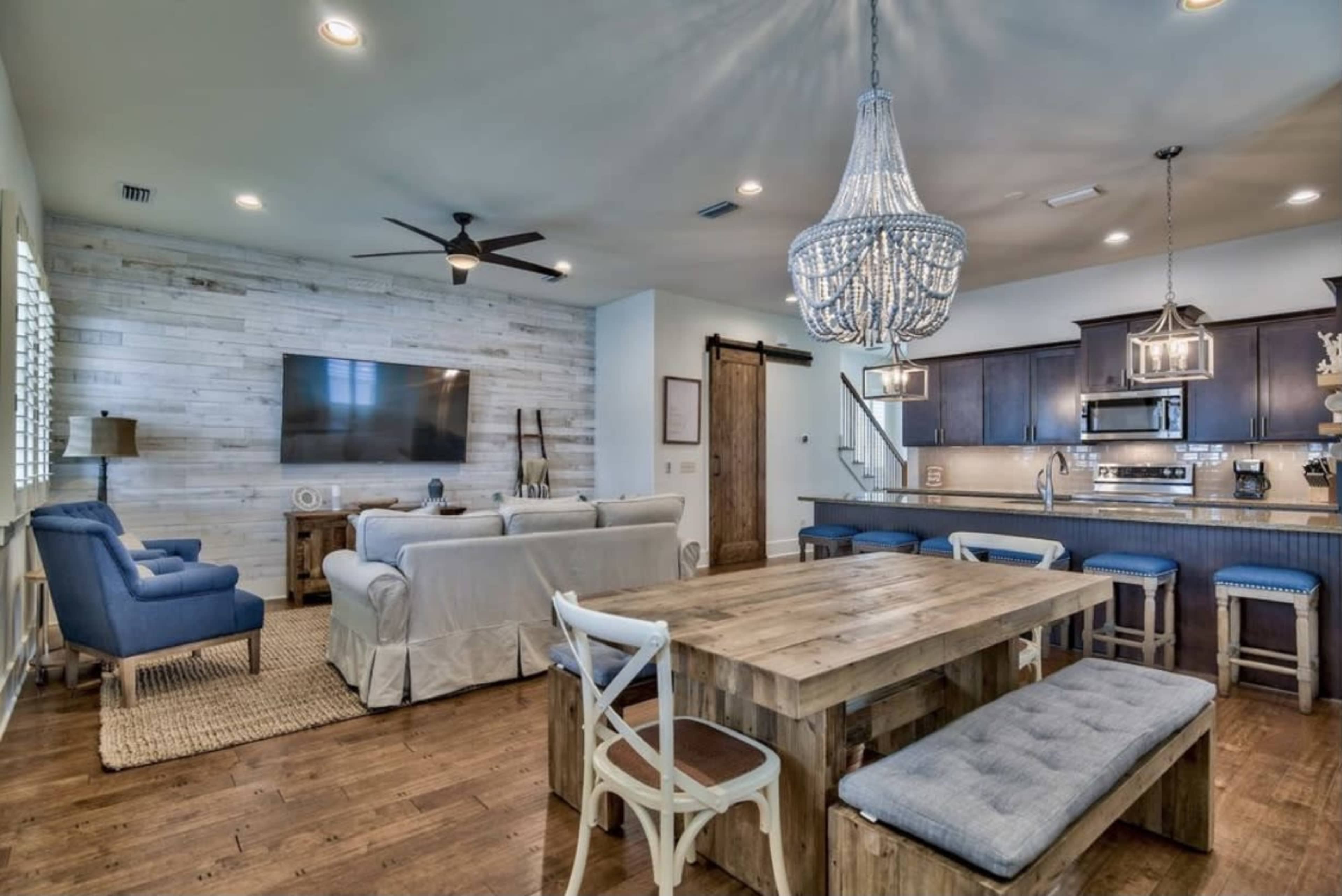 A modern living space featuring a dining table with seating, a stylish chandelier, a large television on a wooden wall, and an open kitchen area.