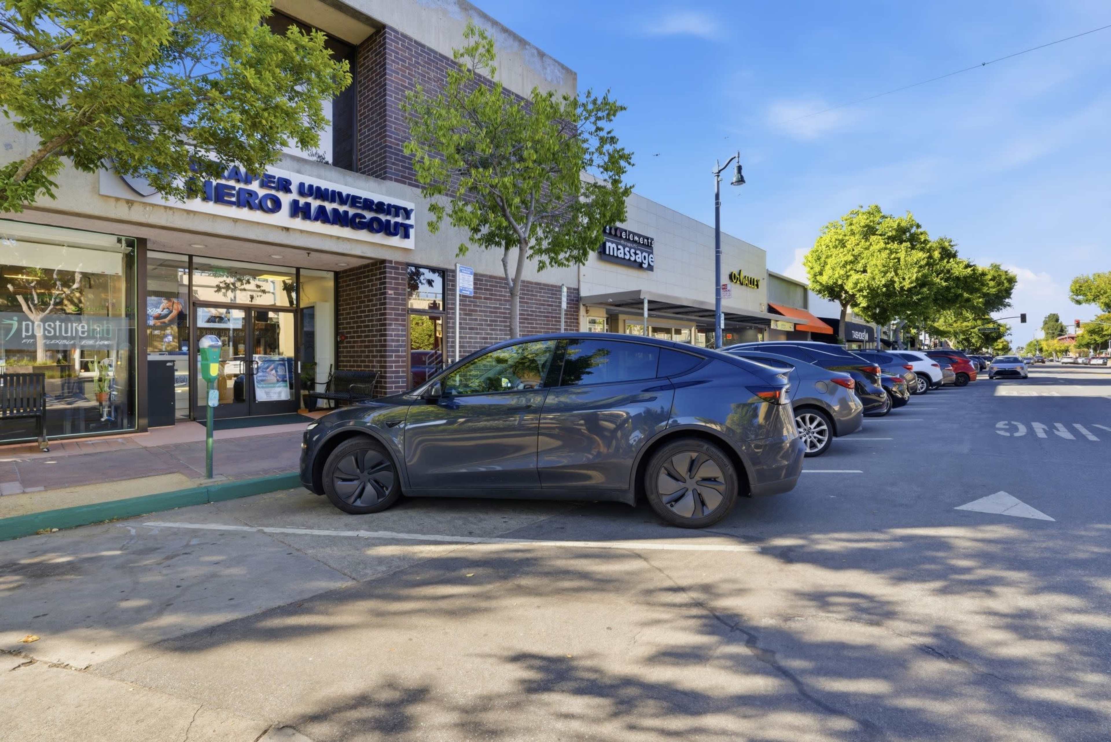 A Tesla car is parked in front of a row of storefronts in a commercial area, with trees lining the street and clear blue skies above.