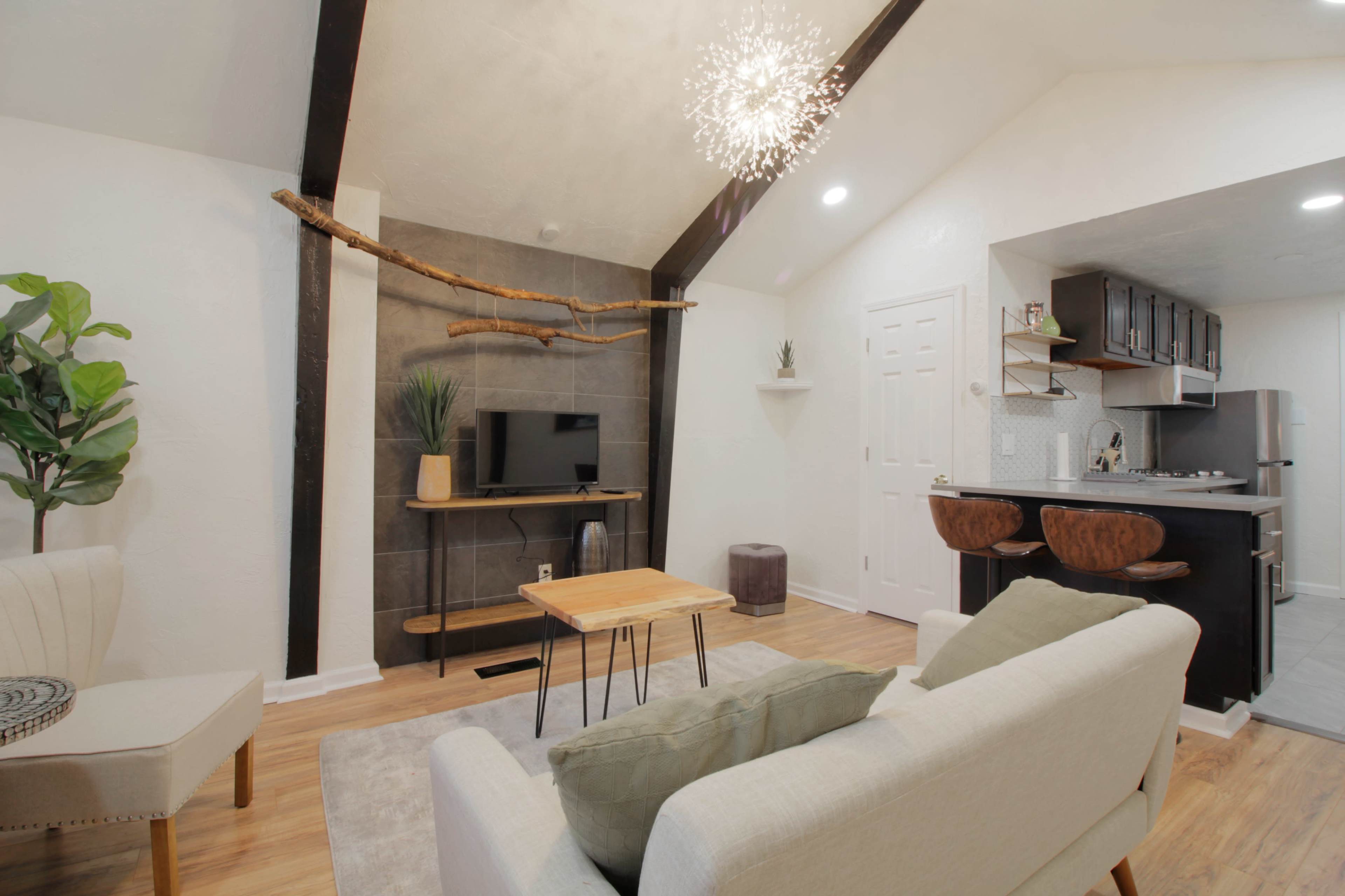A modern living space featuring a light-colored sofa, a wooden coffee table, a TV mounted on a textured wall, and a small kitchen area in the background.
