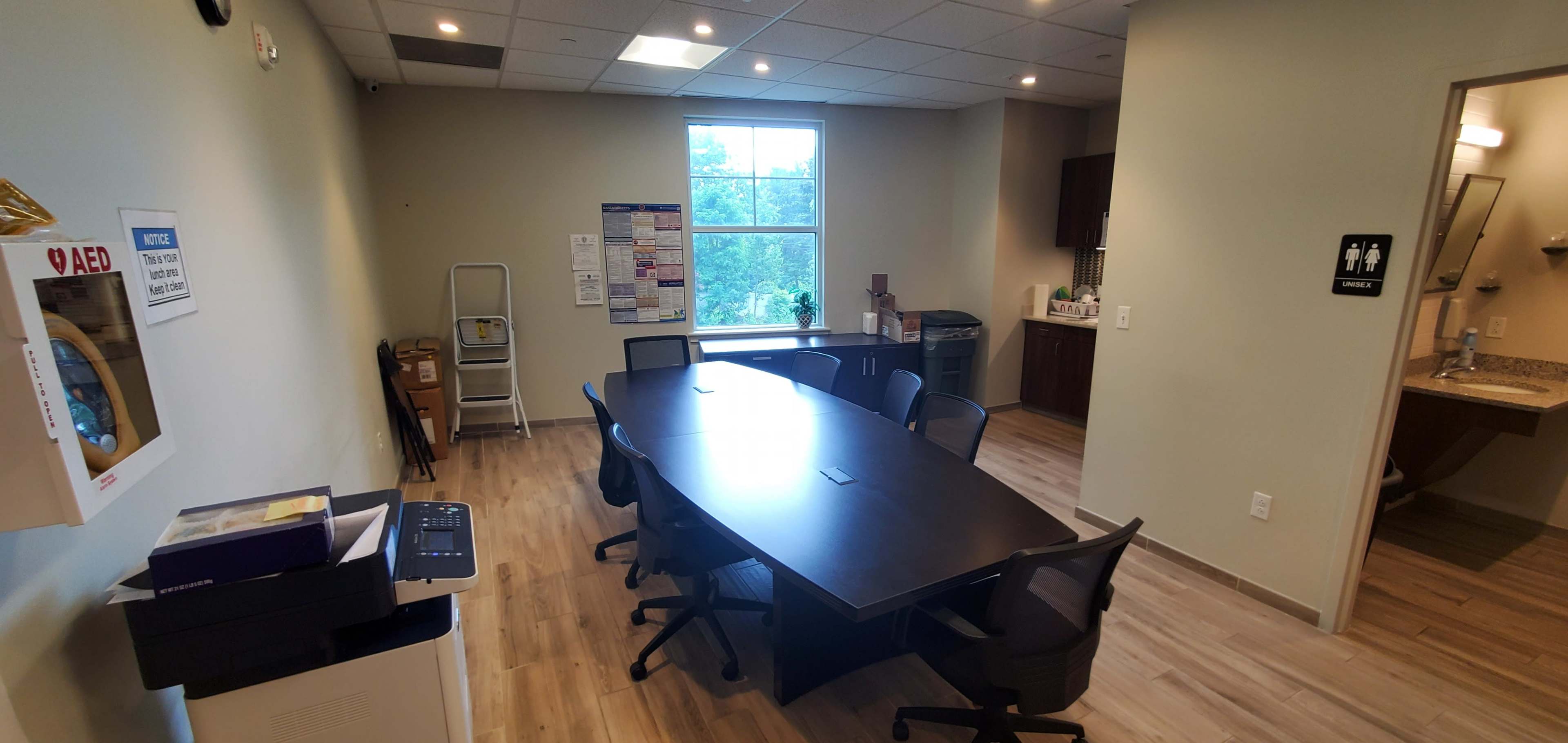 A conference room with a large table surrounded by several black chairs, adjacent to a kitchenette area and a restroom sign.
