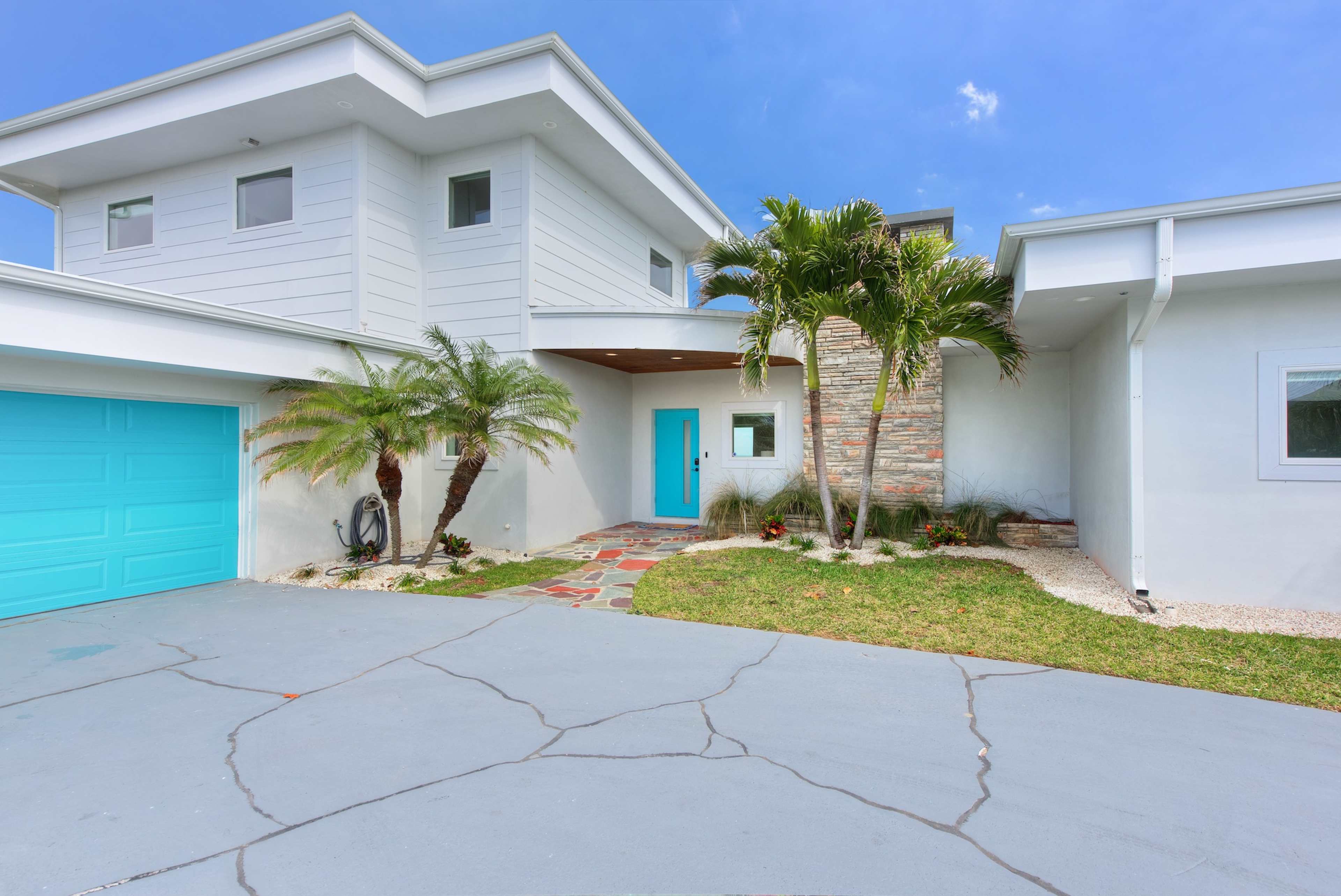 A modern two-story house with a turquoise garage door and a landscaped front yard featuring palm trees and stone pathways.