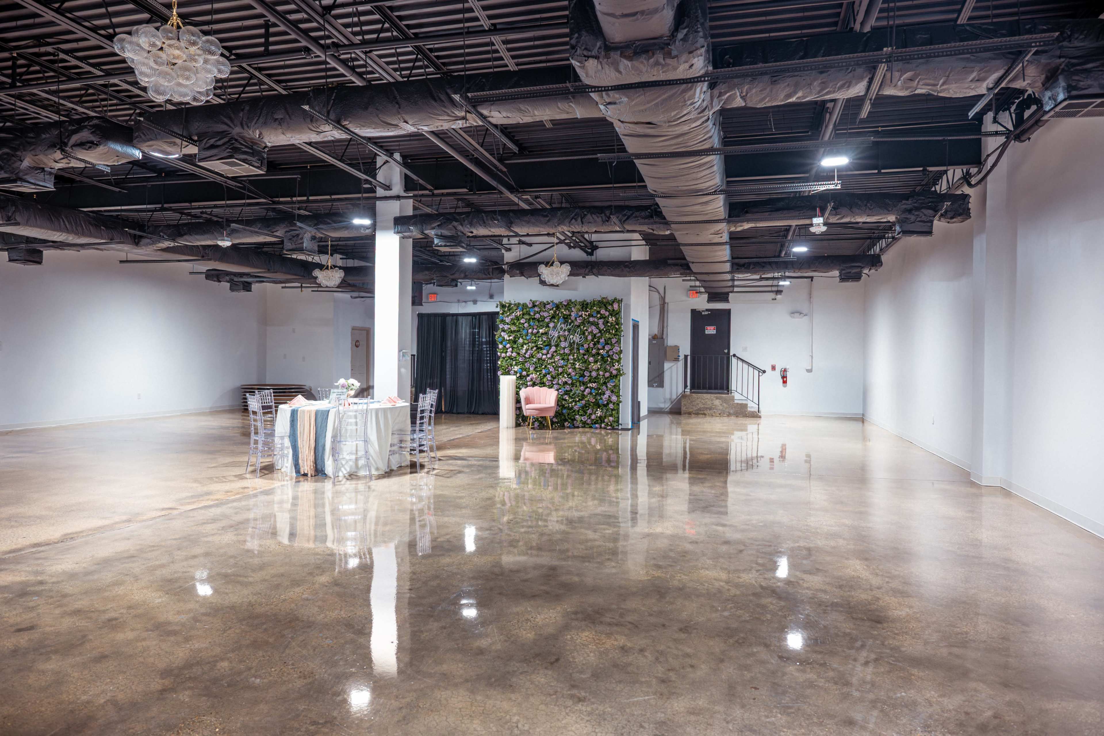 The image shows a spacious, empty event venue with polished concrete floors and a wall featuring greenery.