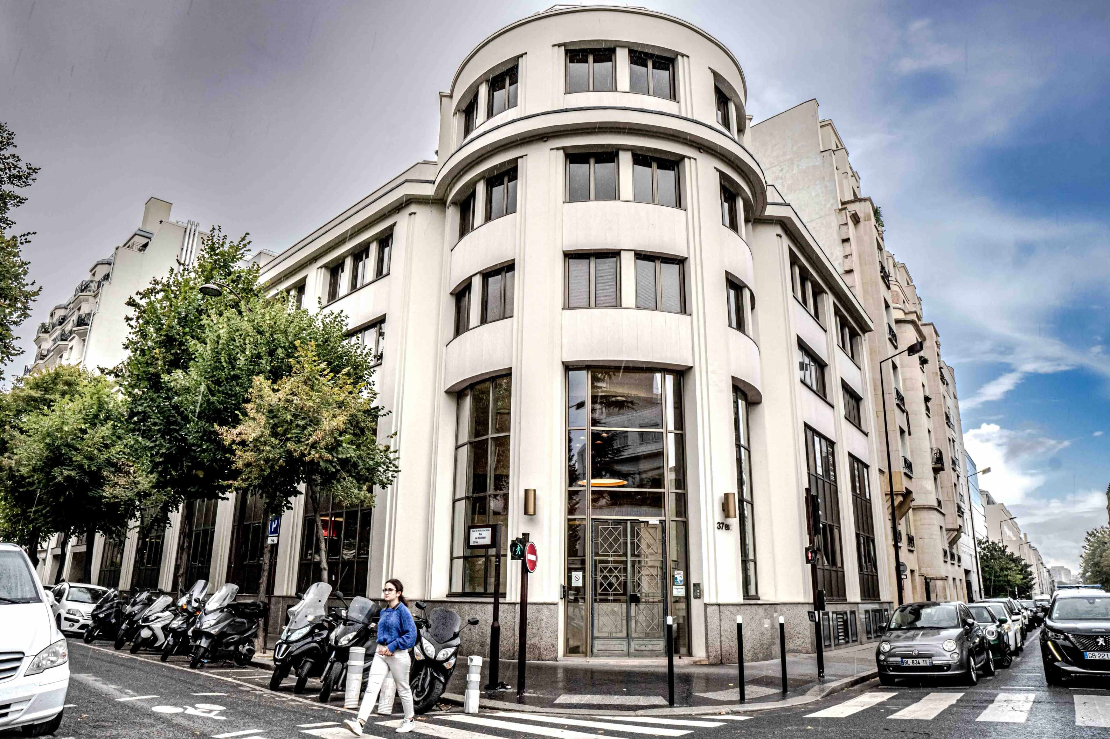 A pedestrian crosses the street in front of a modern building with a distinctive curved façade, surrounded by parked motorcycles and cars.