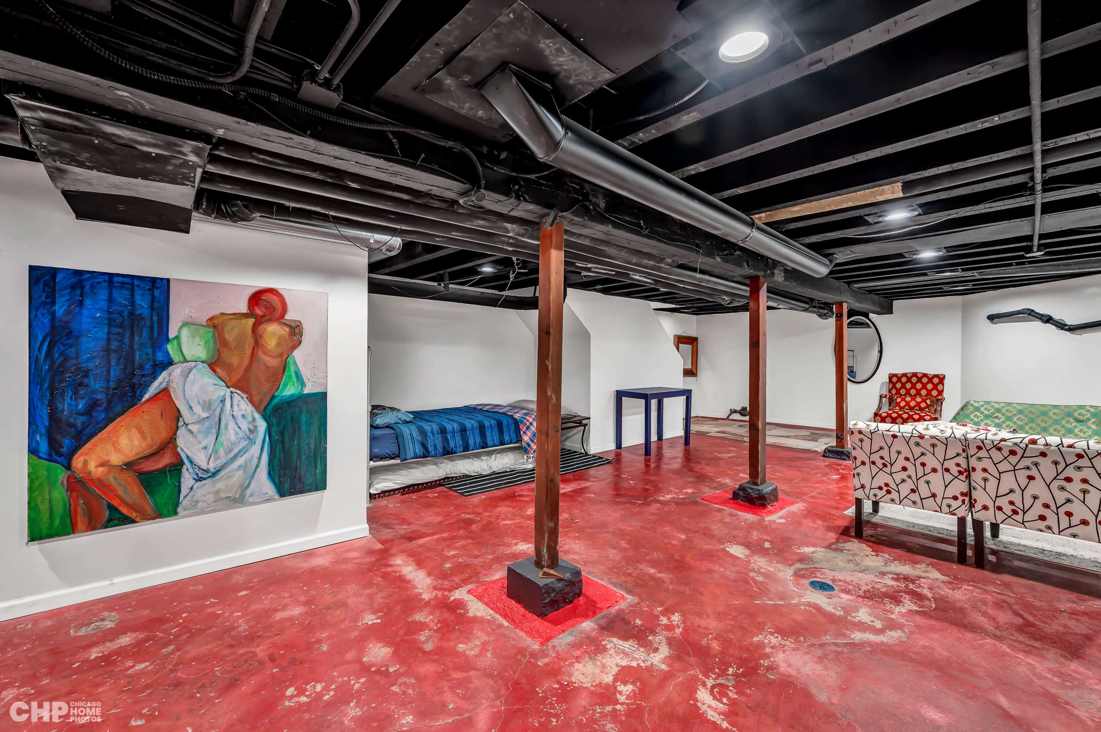 A finished basement with concrete flooring, wooden support beams, a colorful painting on the wall, a bed, and a table set with patterned fabric.