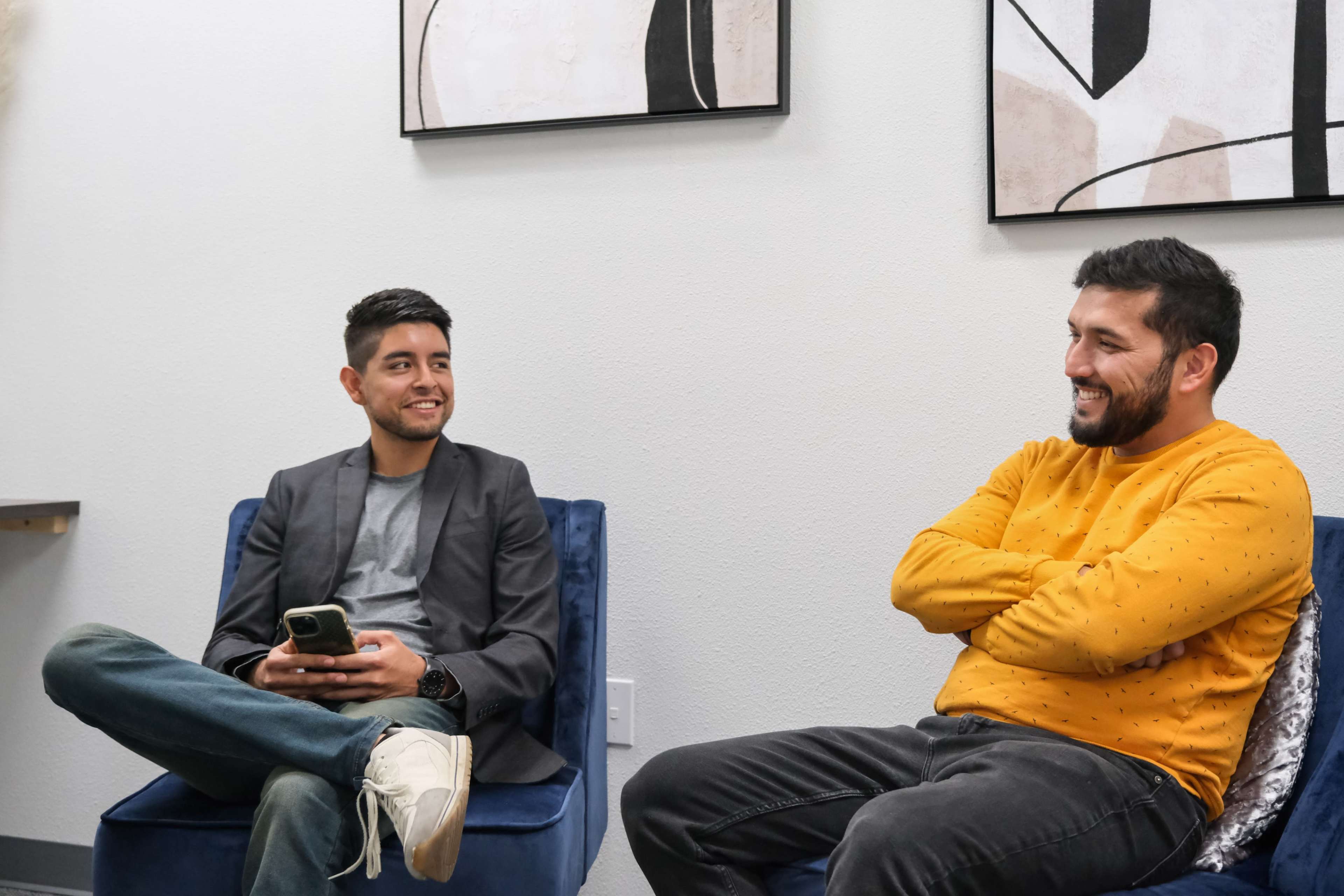 Two men are seated on blue chairs, engaging in conversation, with abstract artwork hanging on the wall behind them.
