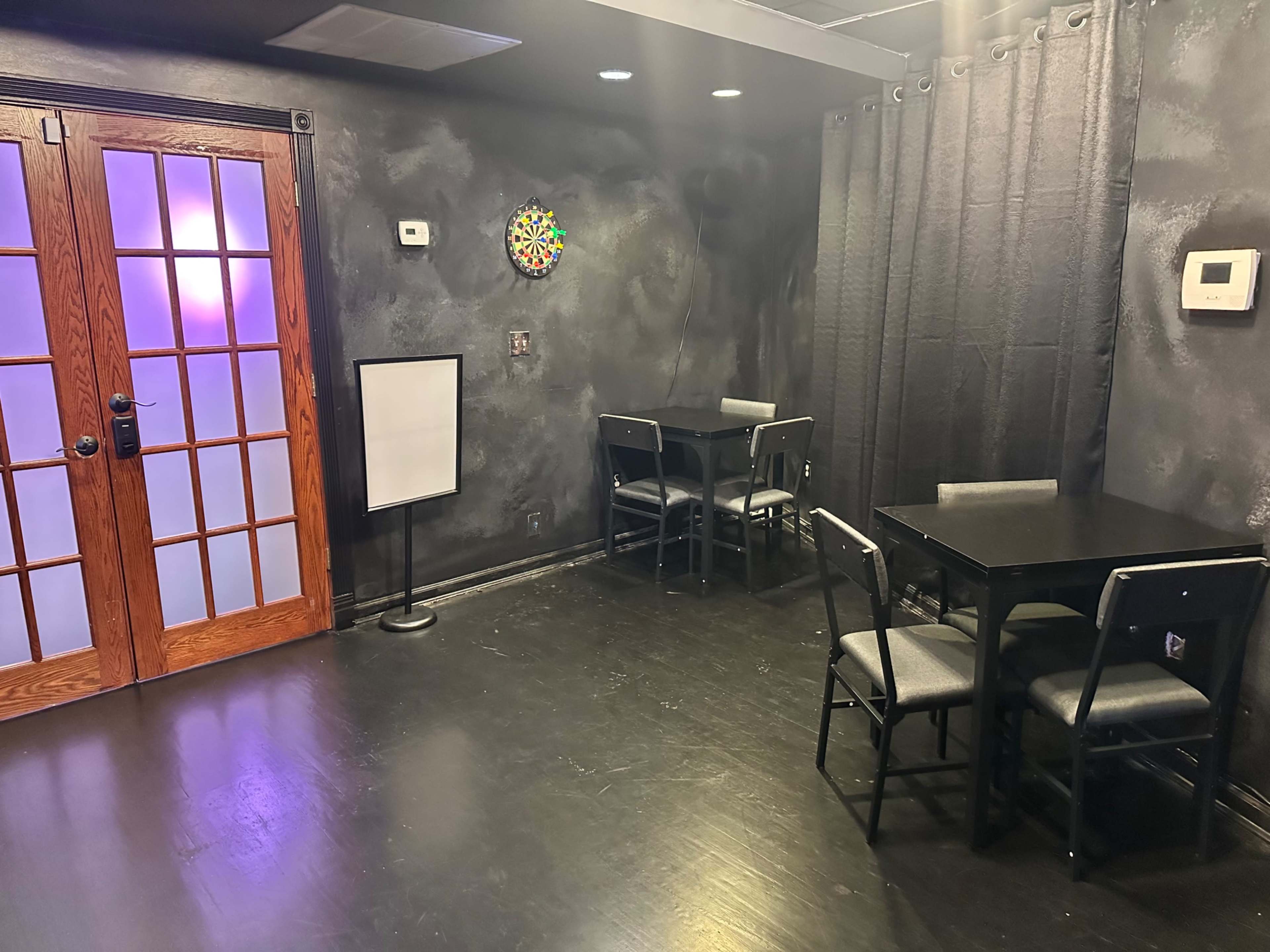 The image shows a dimly lit room with black walls, two black tables with chairs, a dartboard on the wall, and double doors made of glass and wood.