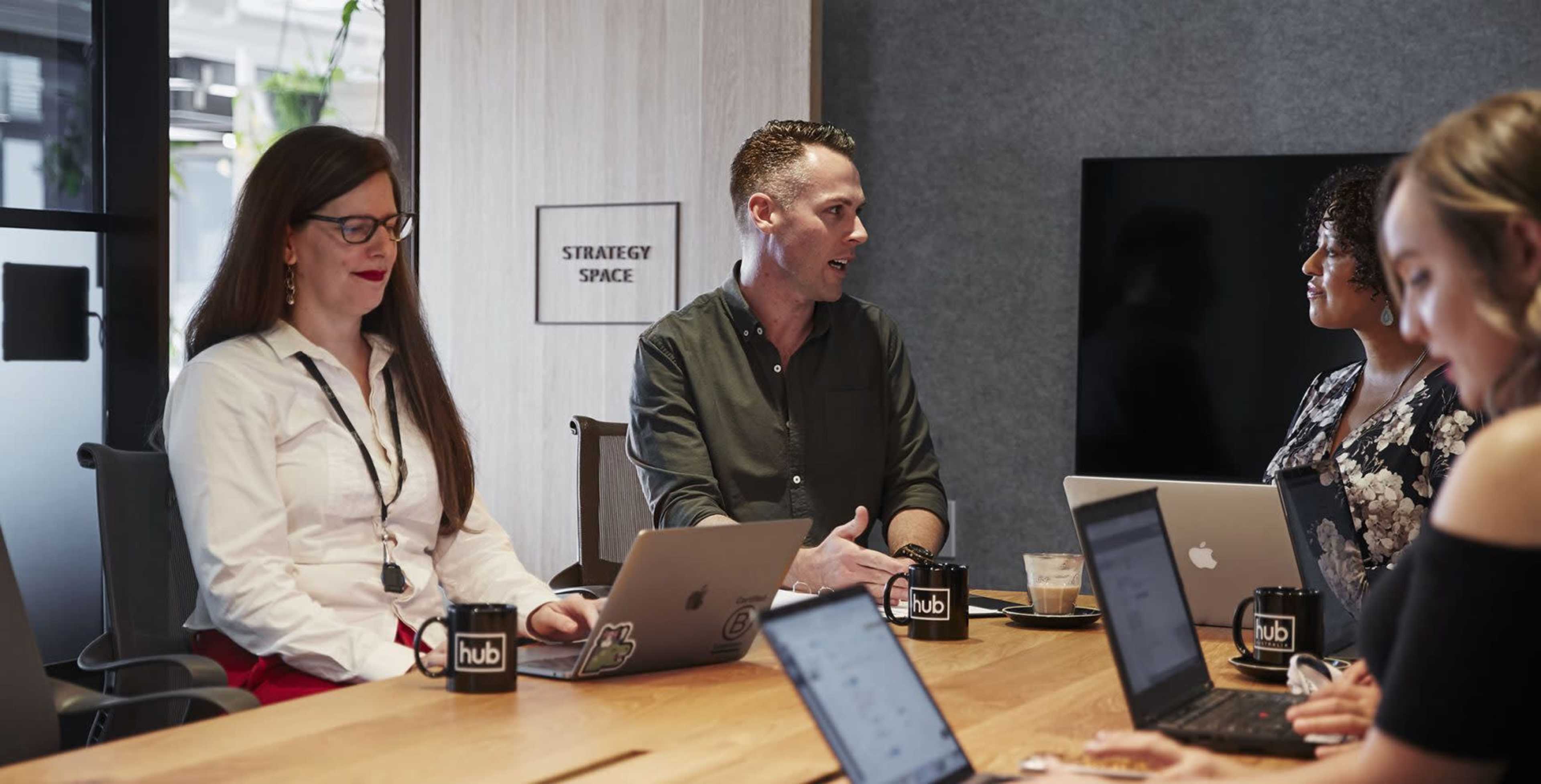 A group of professionals sit around a conference table, engaged in discussion with laptops open in front of them.