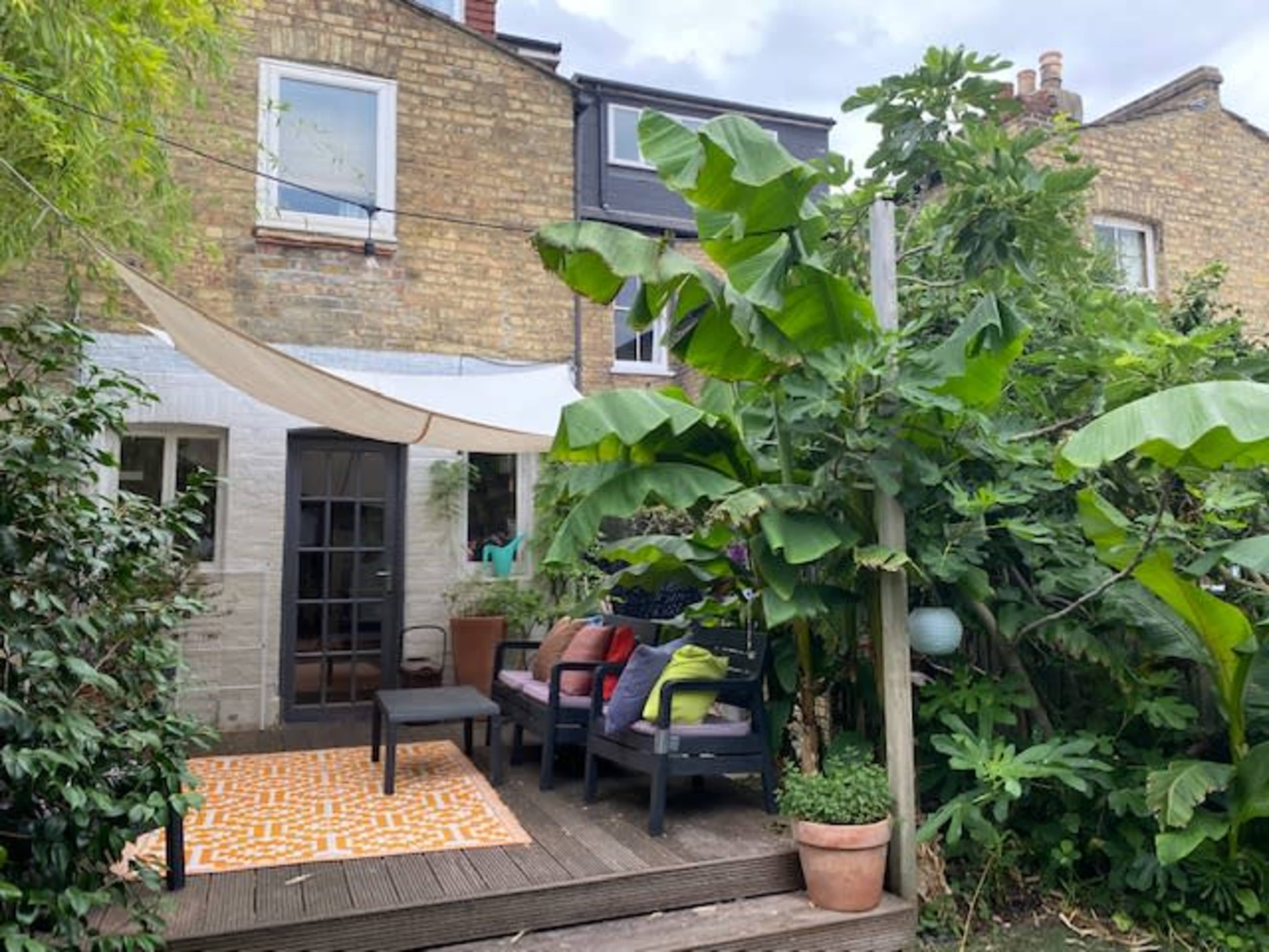 A cozy outdoor seating area with a patterned rug and a backdrop of lush greenery is situated against a brick wall of a house.