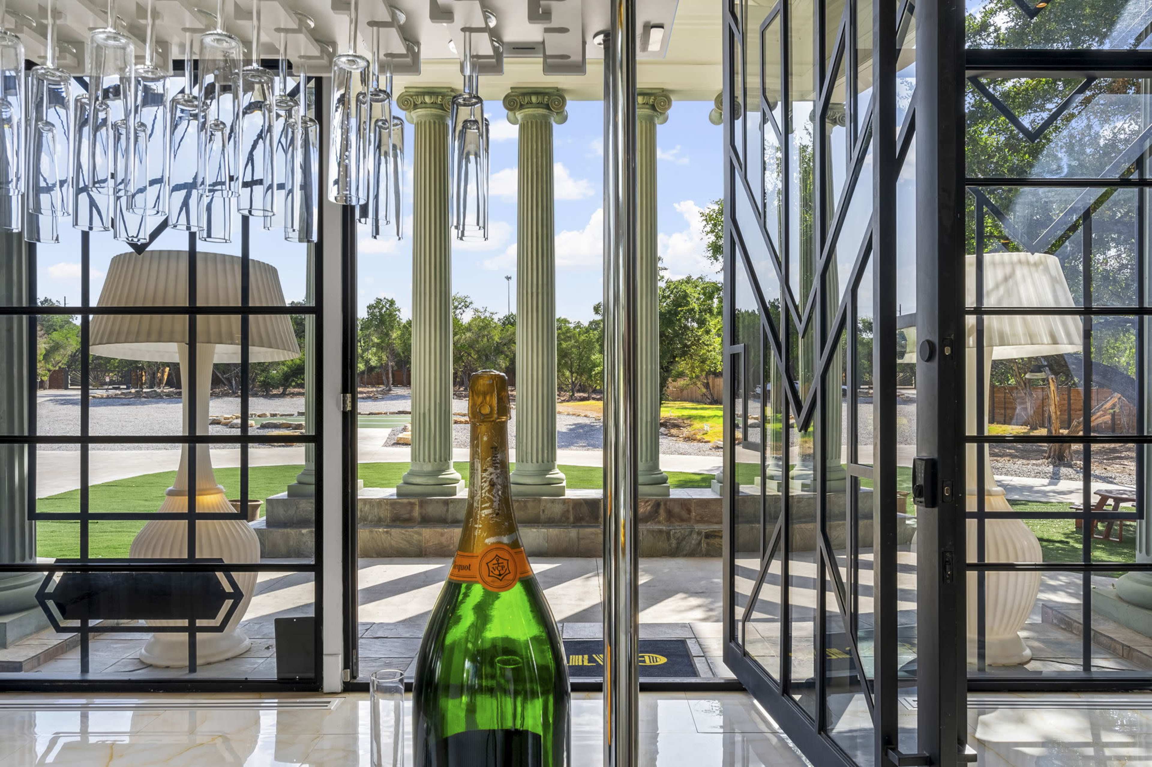 A spacious room with large glass doors opening to a view of pillars and a landscaped area outside, featuring a bottle of champagne in the foreground.