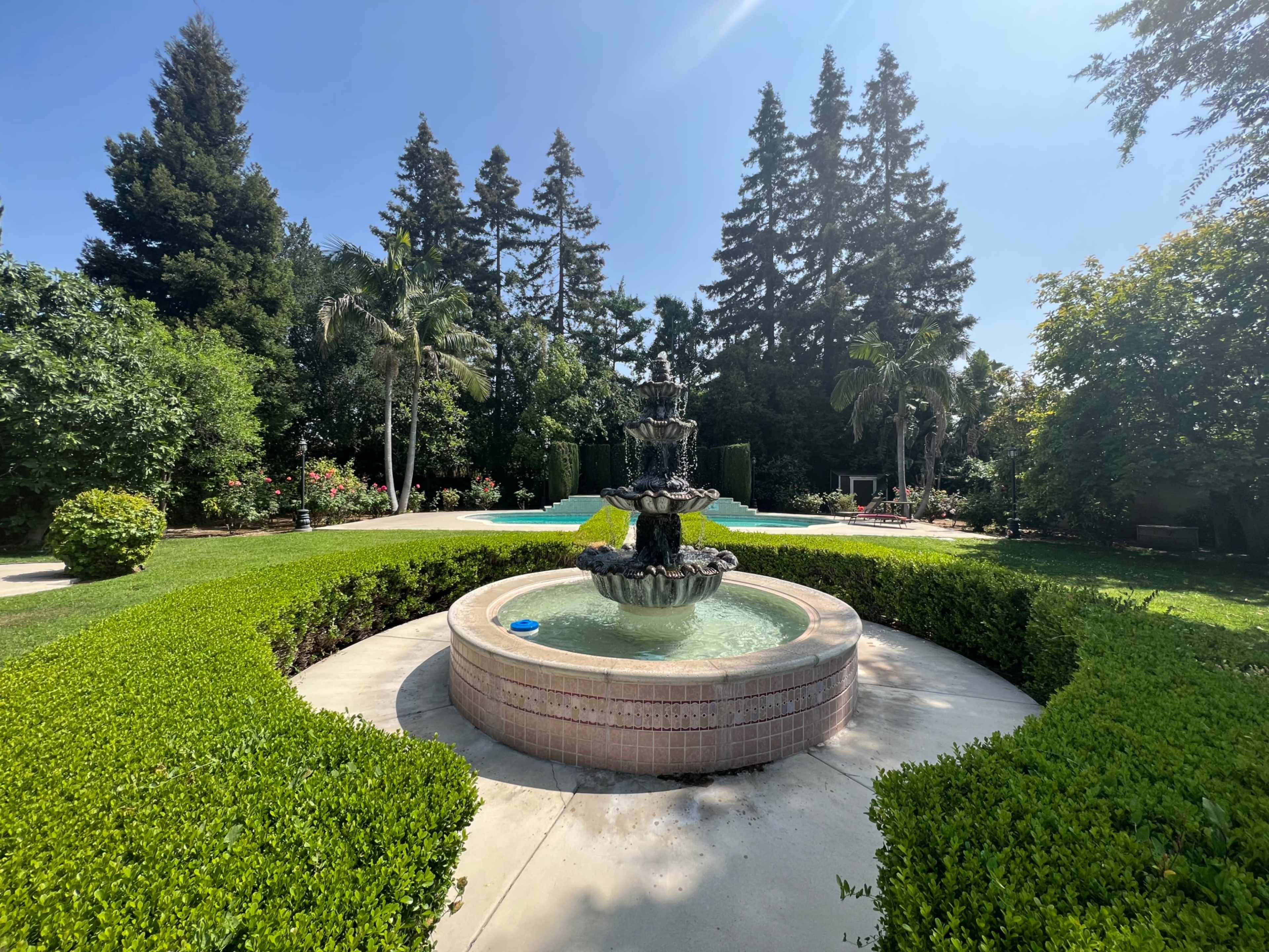 A central fountain surrounded by neatly trimmed hedges is located in a landscaped garden with trees and a swimming pool in the background.