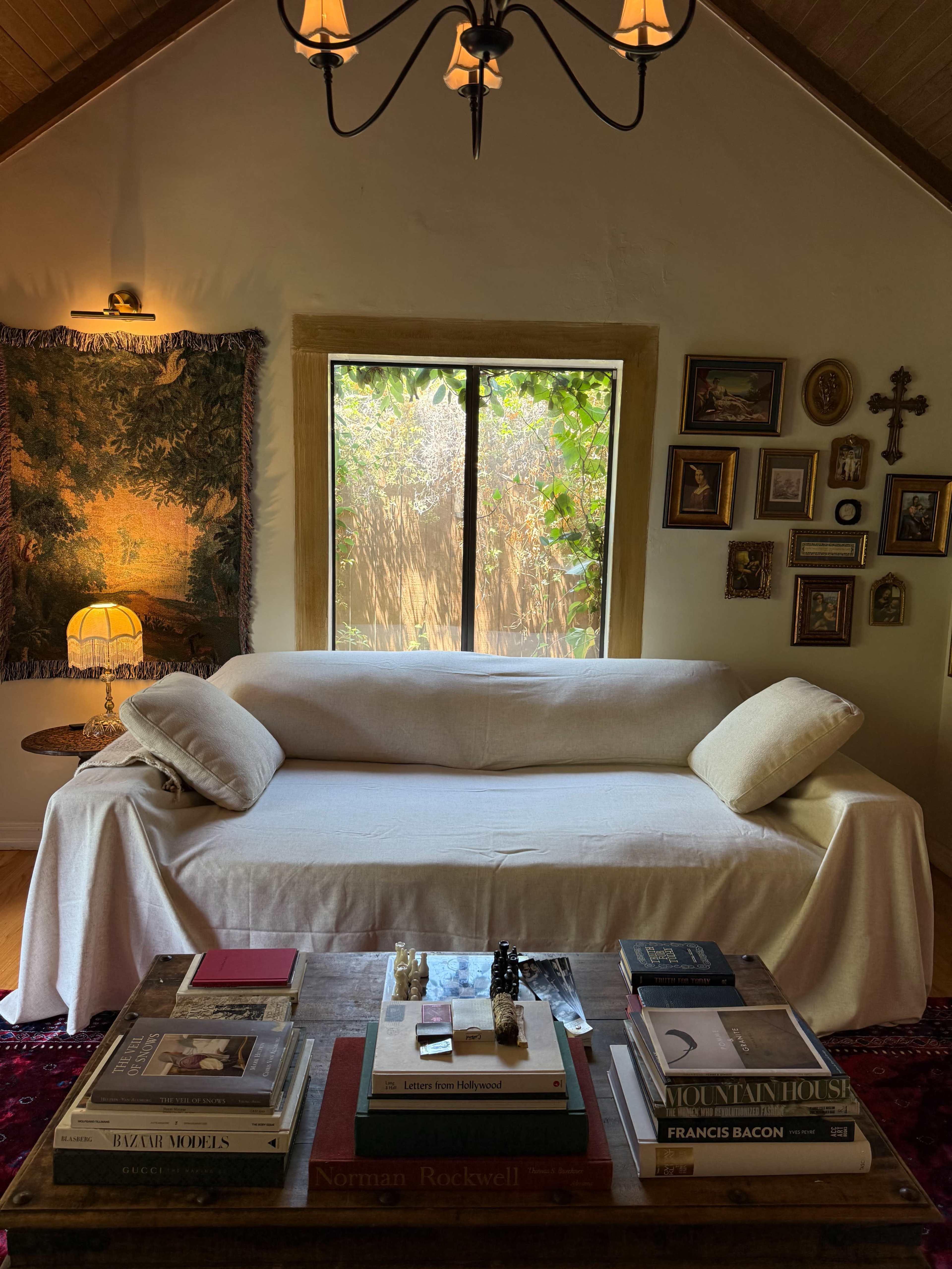 A white sofa is positioned in a cozy living room with a window displaying greenery, surrounded by a variety of framed artworks and books on a wooden coffee table.
