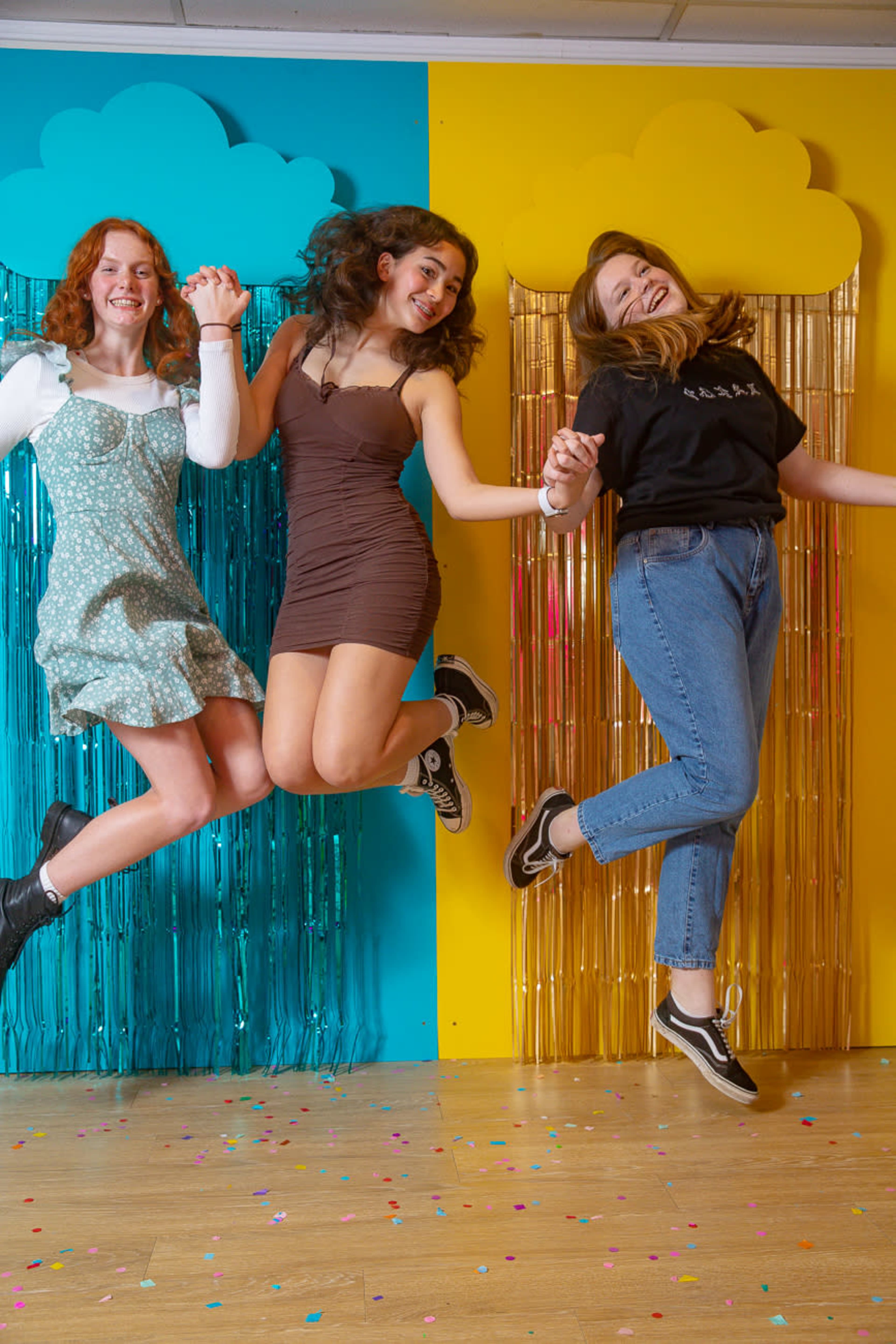Three young women jump in unison in front of colorful backdrop with fringed decorations and scattered confetti on the ground.