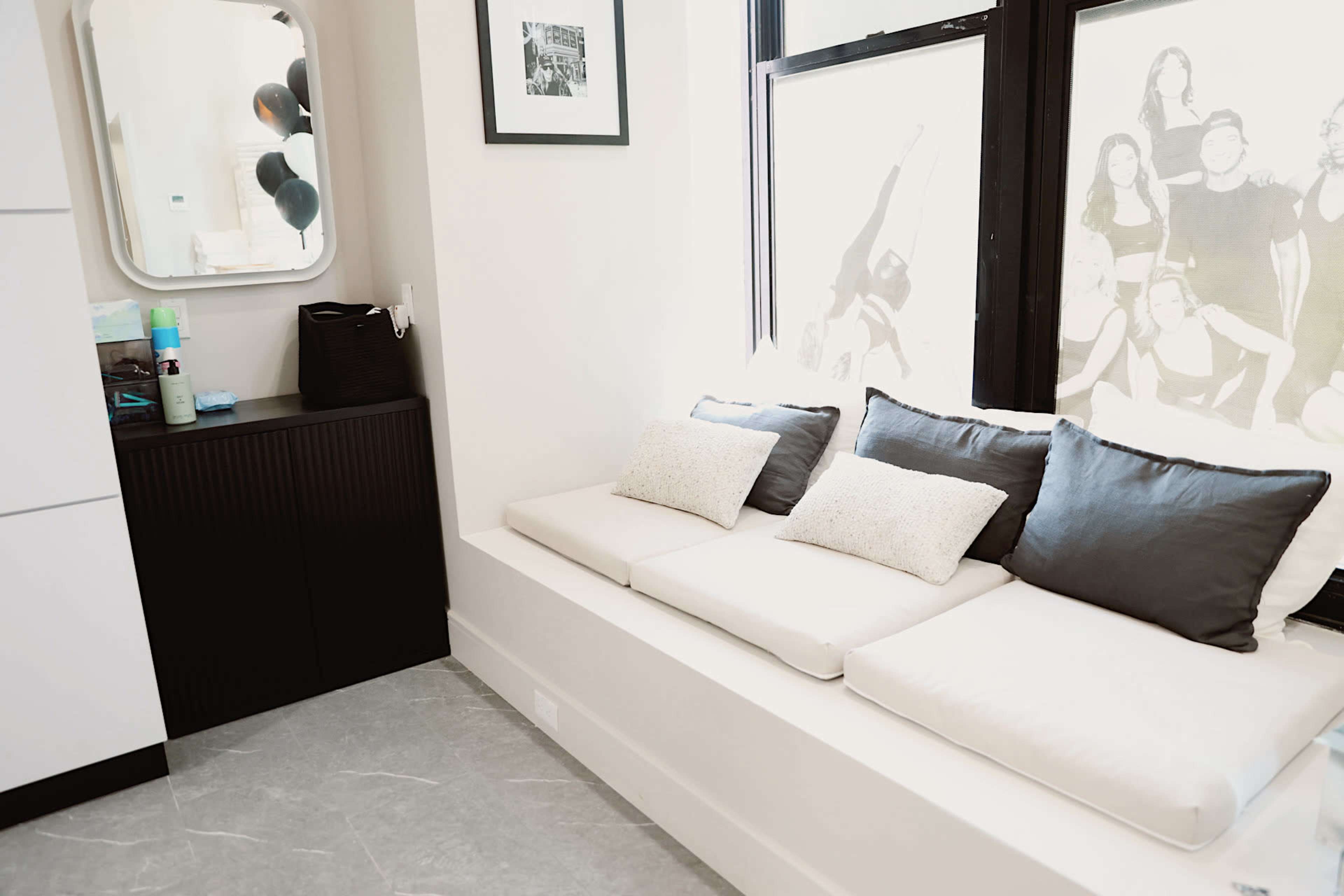 The image shows a modern, minimalistic interior featuring a cushioned seating area beside a large window, a mirror, and a dark cabinet.