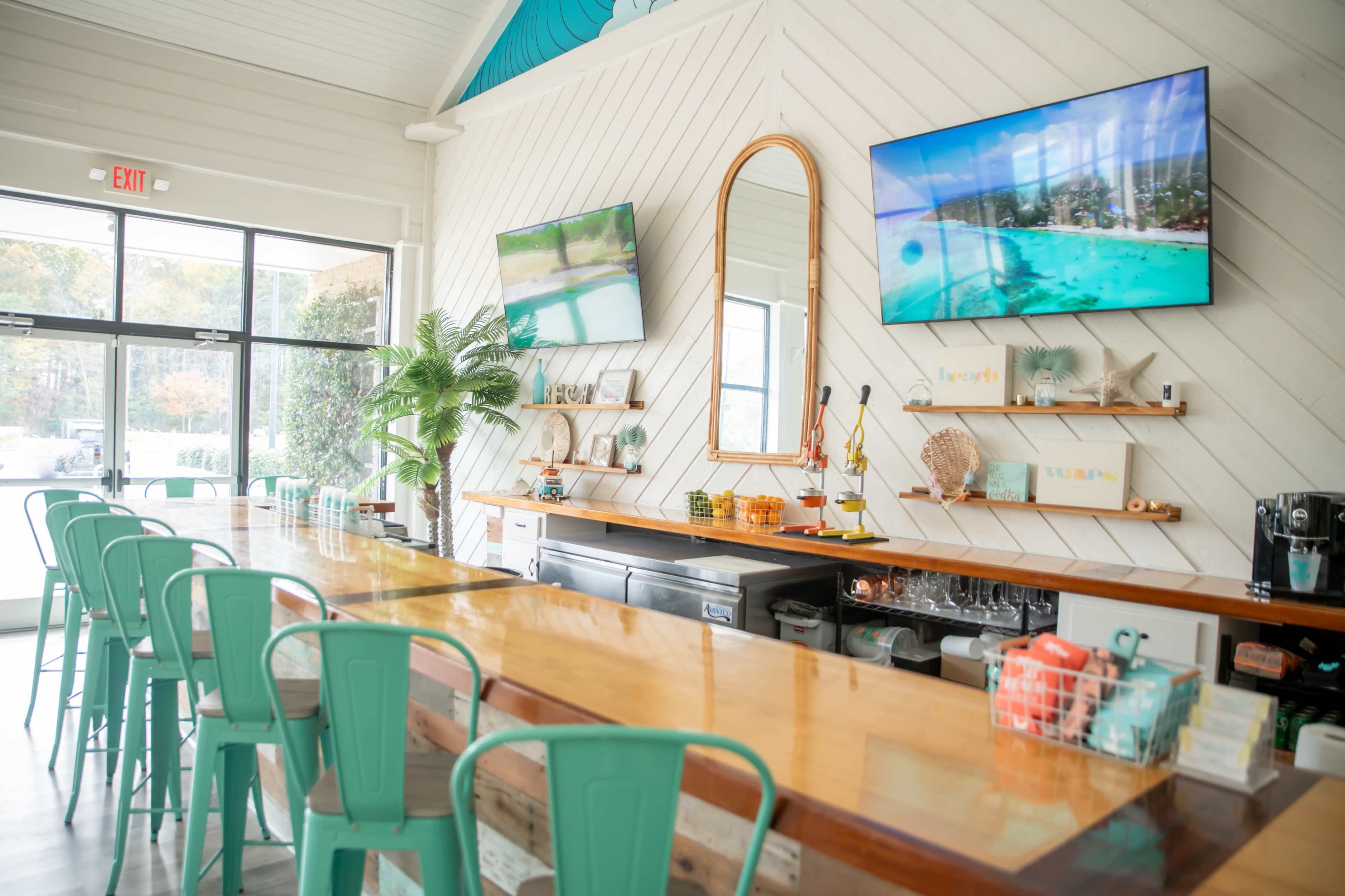 The image depicts a light-filled bar area with a wooden countertop, green metal chairs, two television screens displaying beach scenes, and shelves decorated with coastal-themed items.