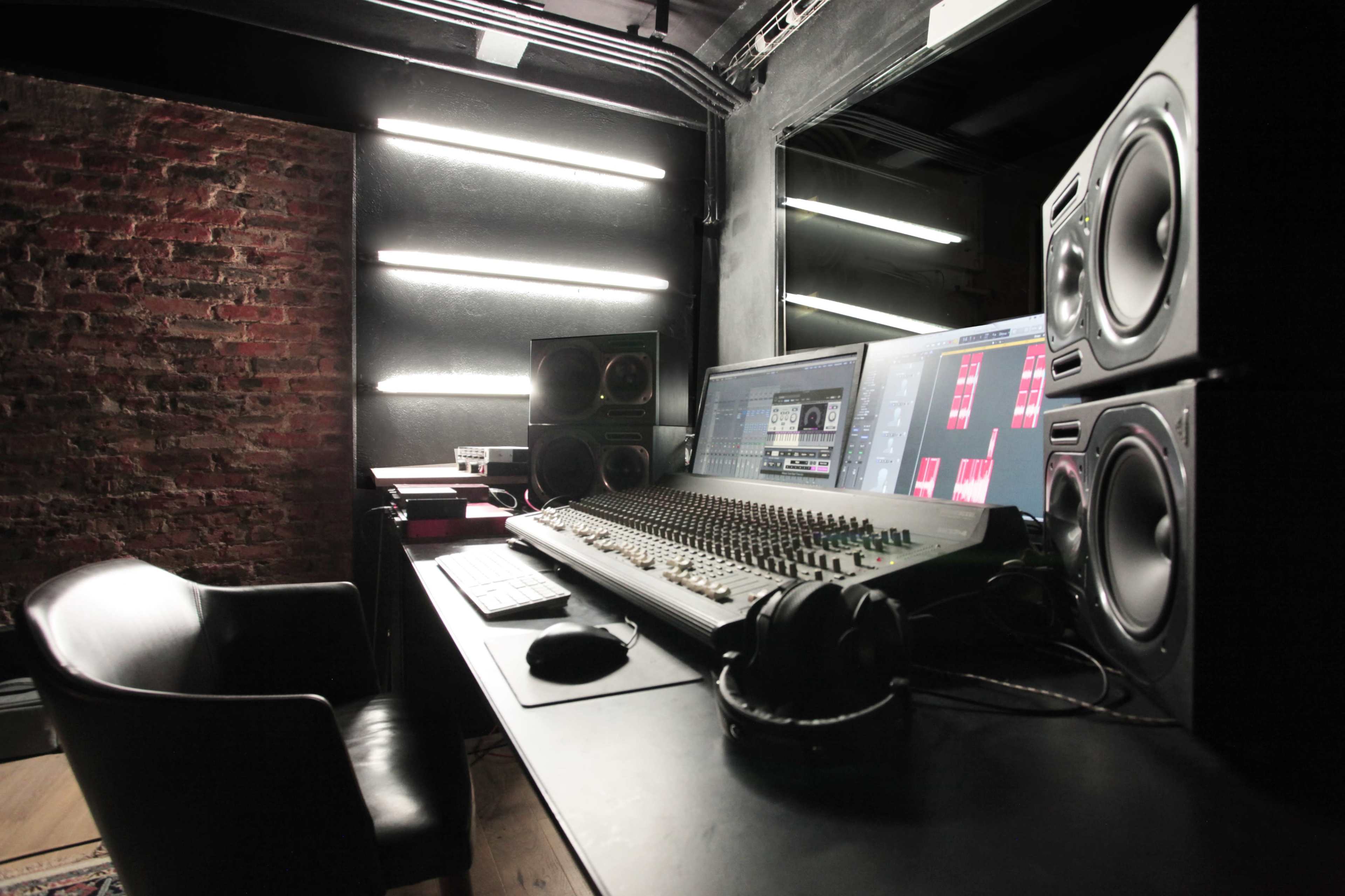 The image shows a modern music studio with a mixing console, studio monitors, and a computer setup against a brick wall.