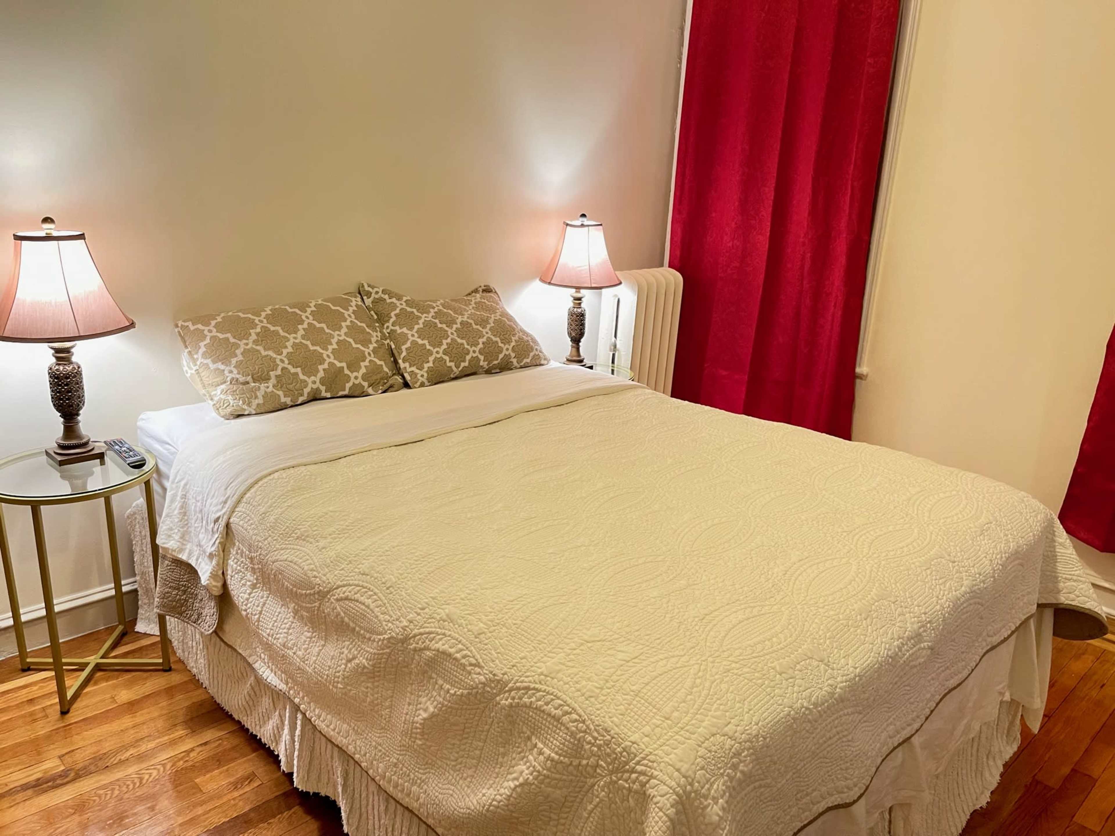 The image shows a neatly made bed with a light-colored quilt, flanked by two lamps, in a room with red curtains and wooden flooring.