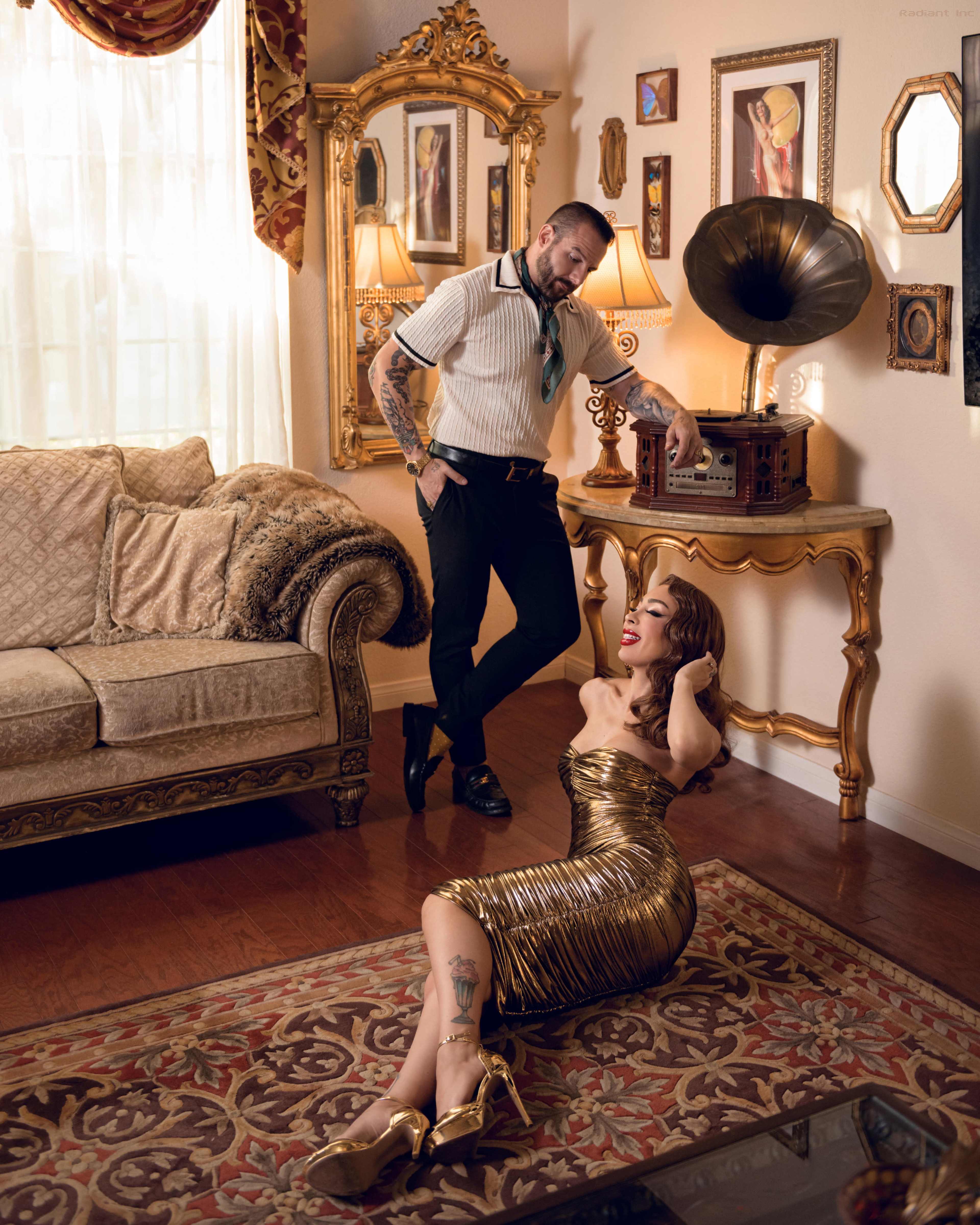 A man stands beside a vintage record player while a woman in a gold dress lounges on the floor of a well-decorated room with rich furnishings.