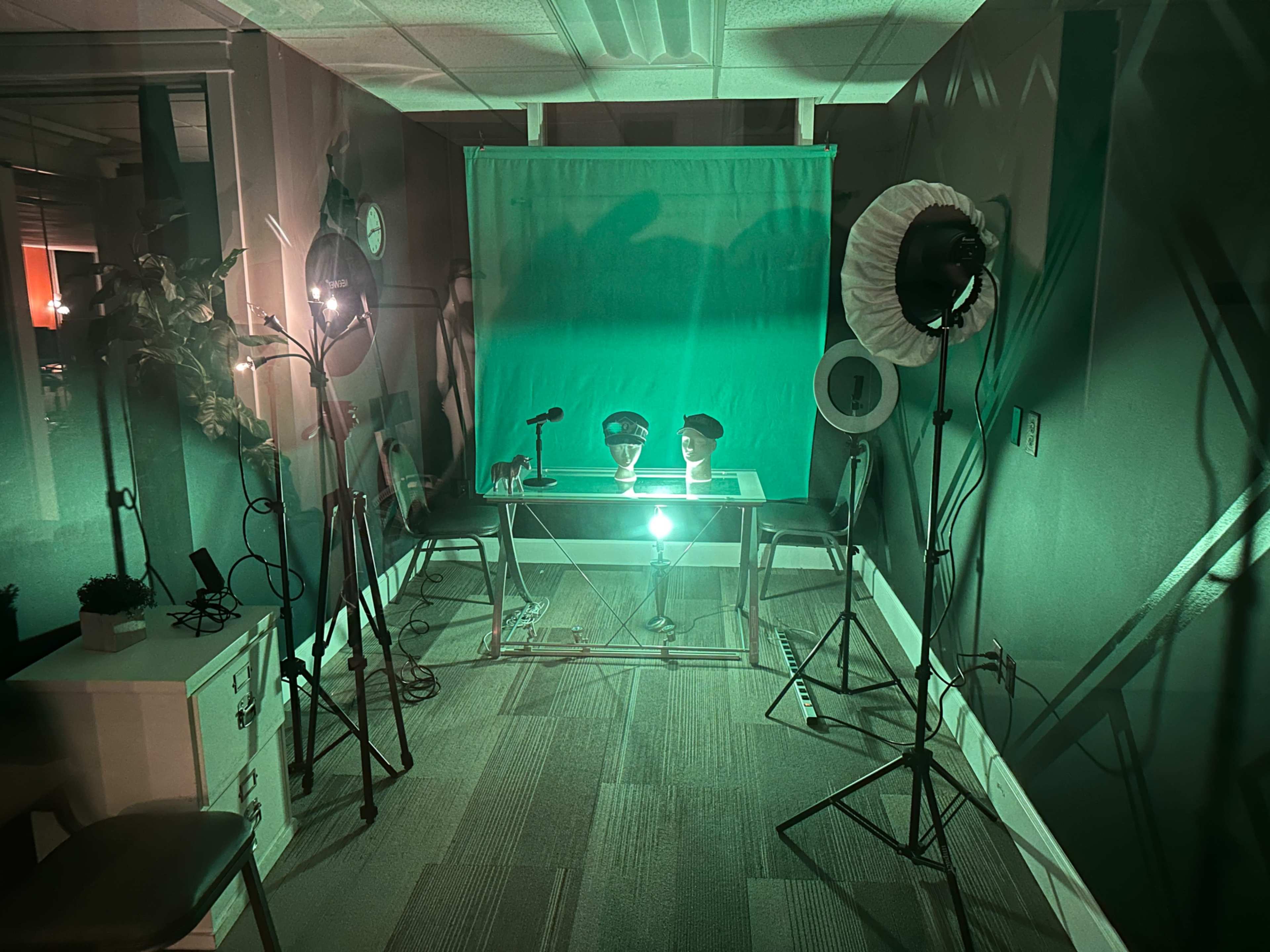The image shows a dimly lit studio setup with a green backdrop, lighting equipment, and a table positioned in the center.