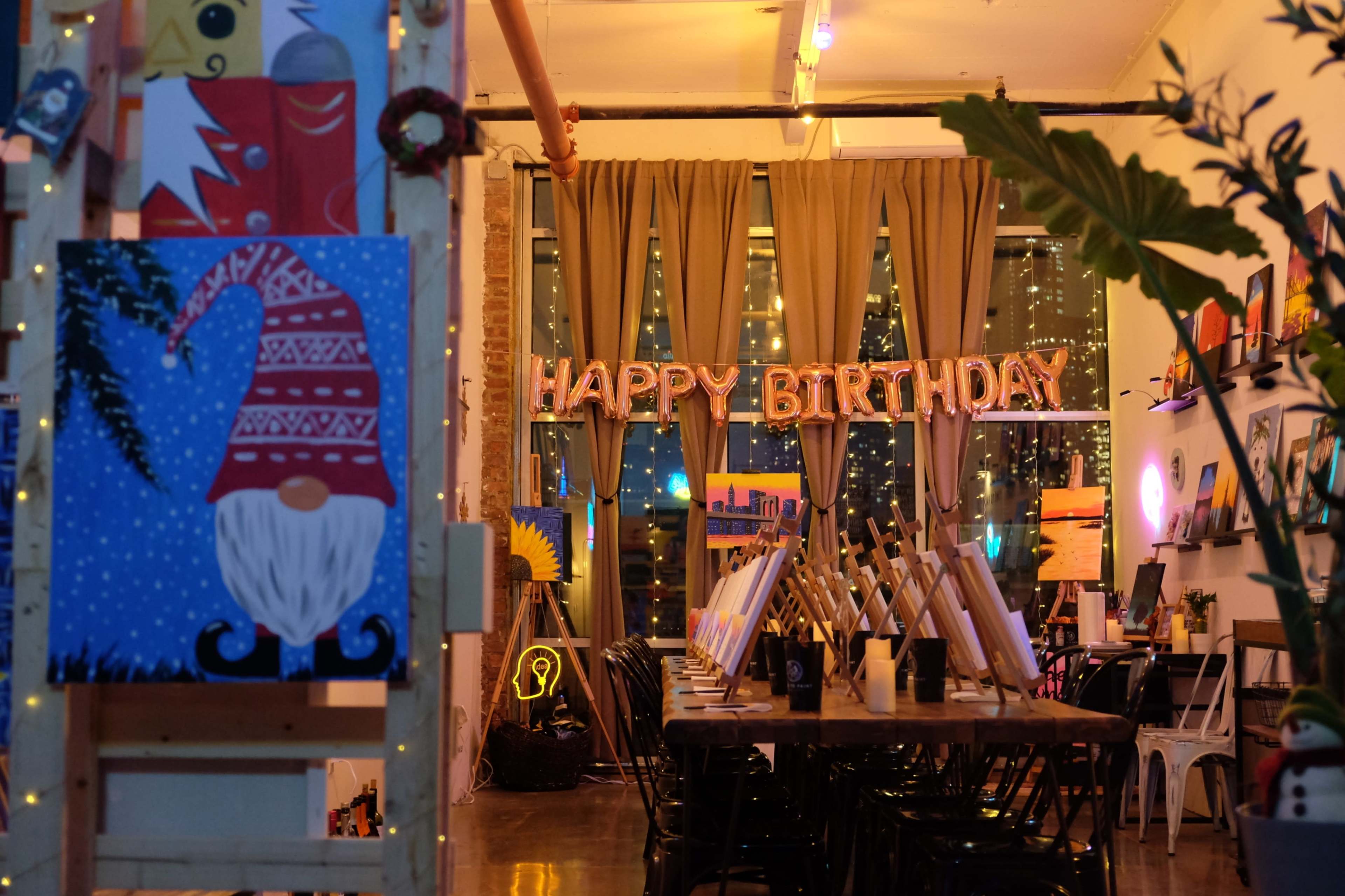 The image shows an art studio decorated for a birthday celebration, featuring a "HAPPY BIRTHDAY" sign, artwork on easels, and ambient lighting.