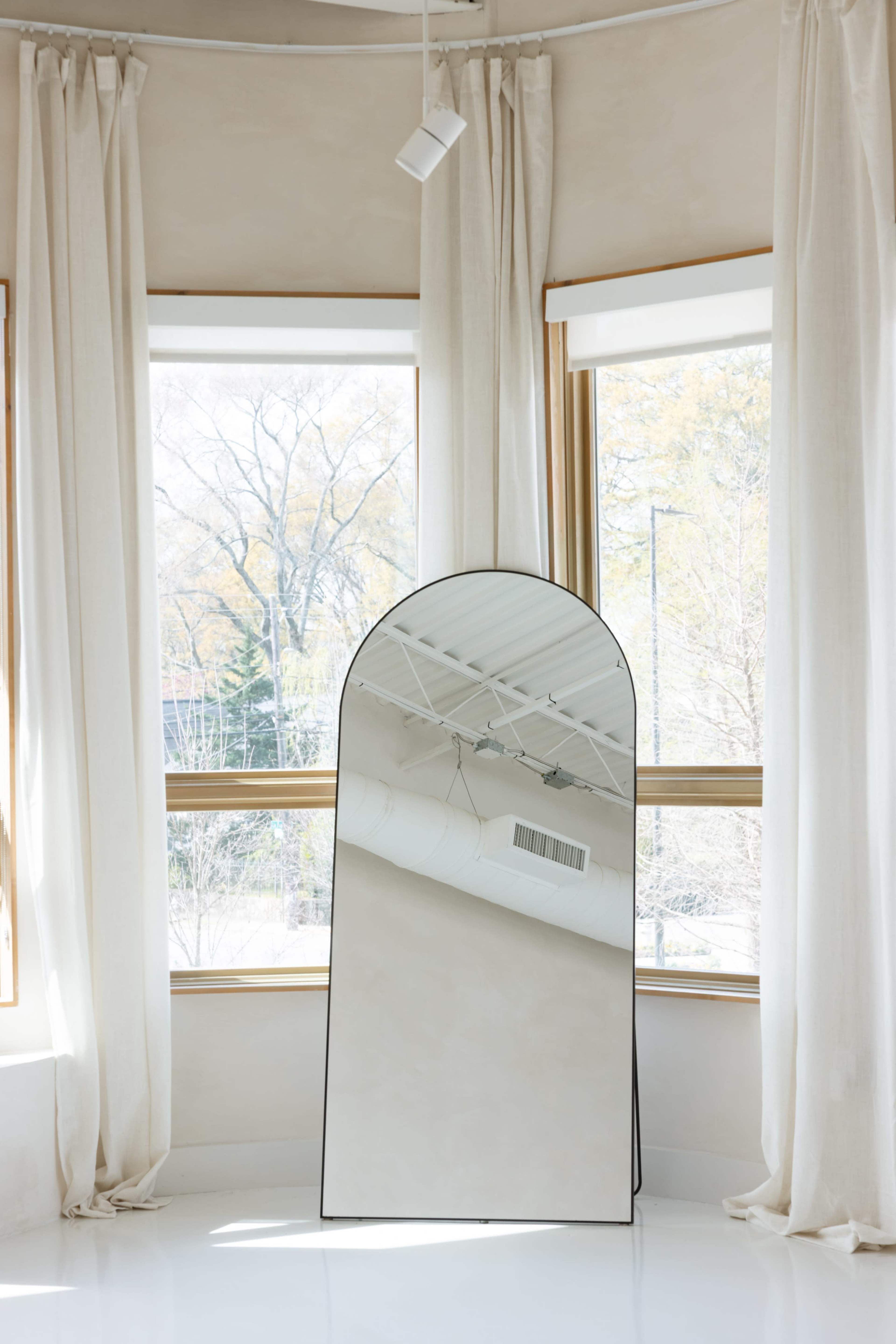 A tall, arched mirror stands in the corner of a room with large windows and sheer white curtains.