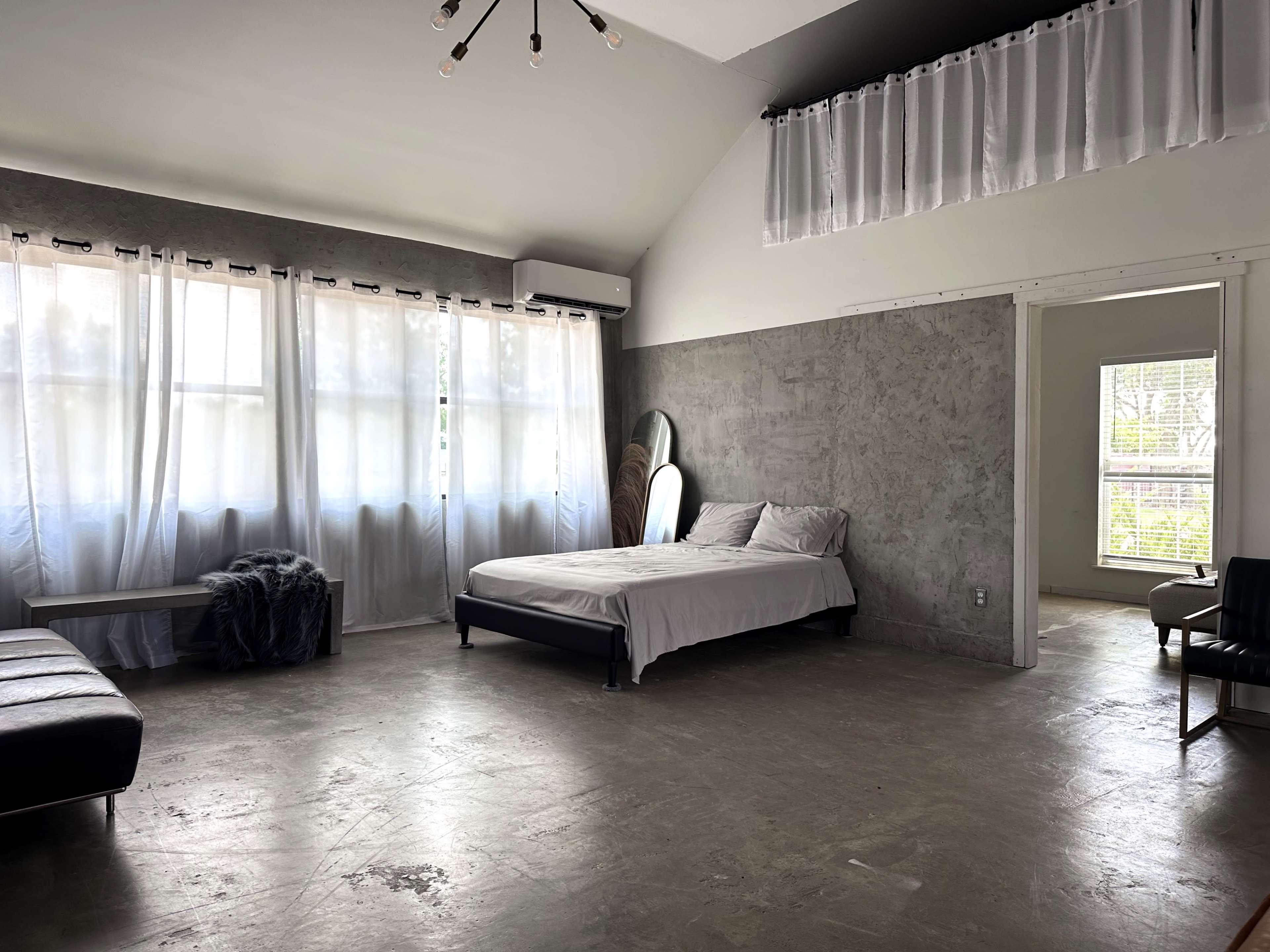 A minimalist bedroom with a bed, a large mirror, a couch, and floor-to-ceiling windows dressed in sheer curtains.