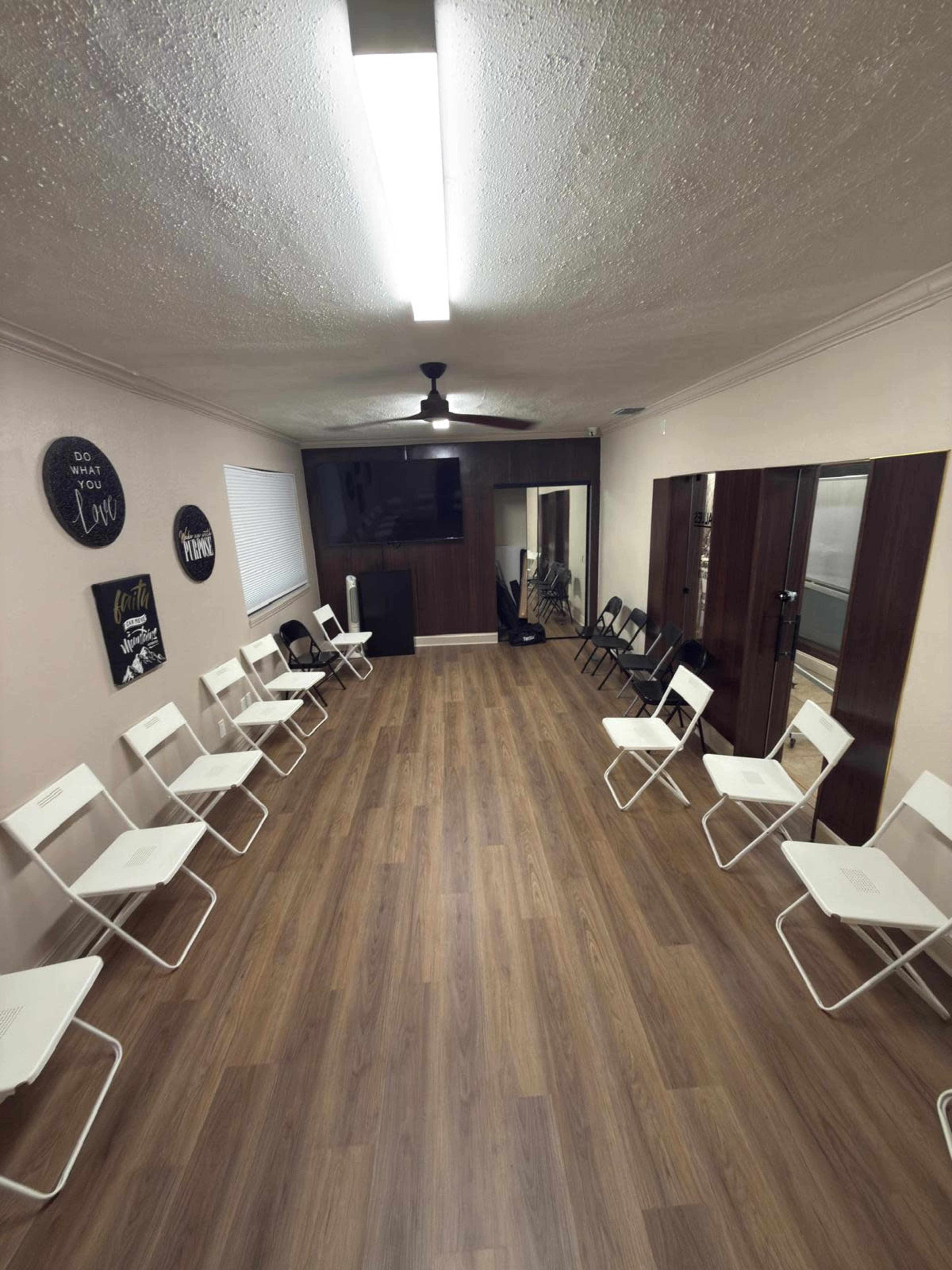 The image shows a room with a light wooden floor, featuring white folding chairs arranged in a circular pattern, and walls adorned with wall art.