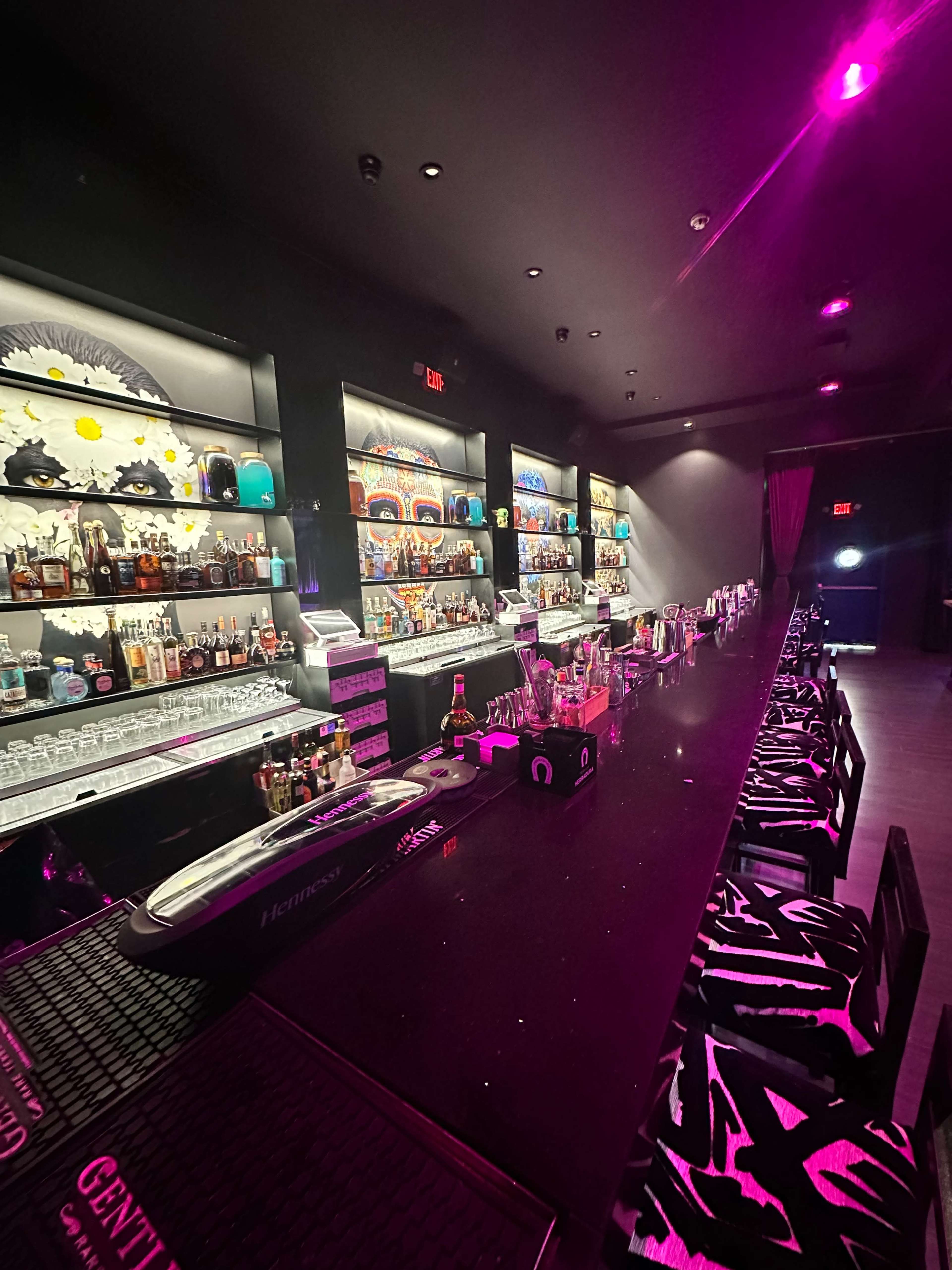 A dimly lit bar with a long black counter and shelves lined with various bottles and colorful decorations in a space illuminated by pink lights.