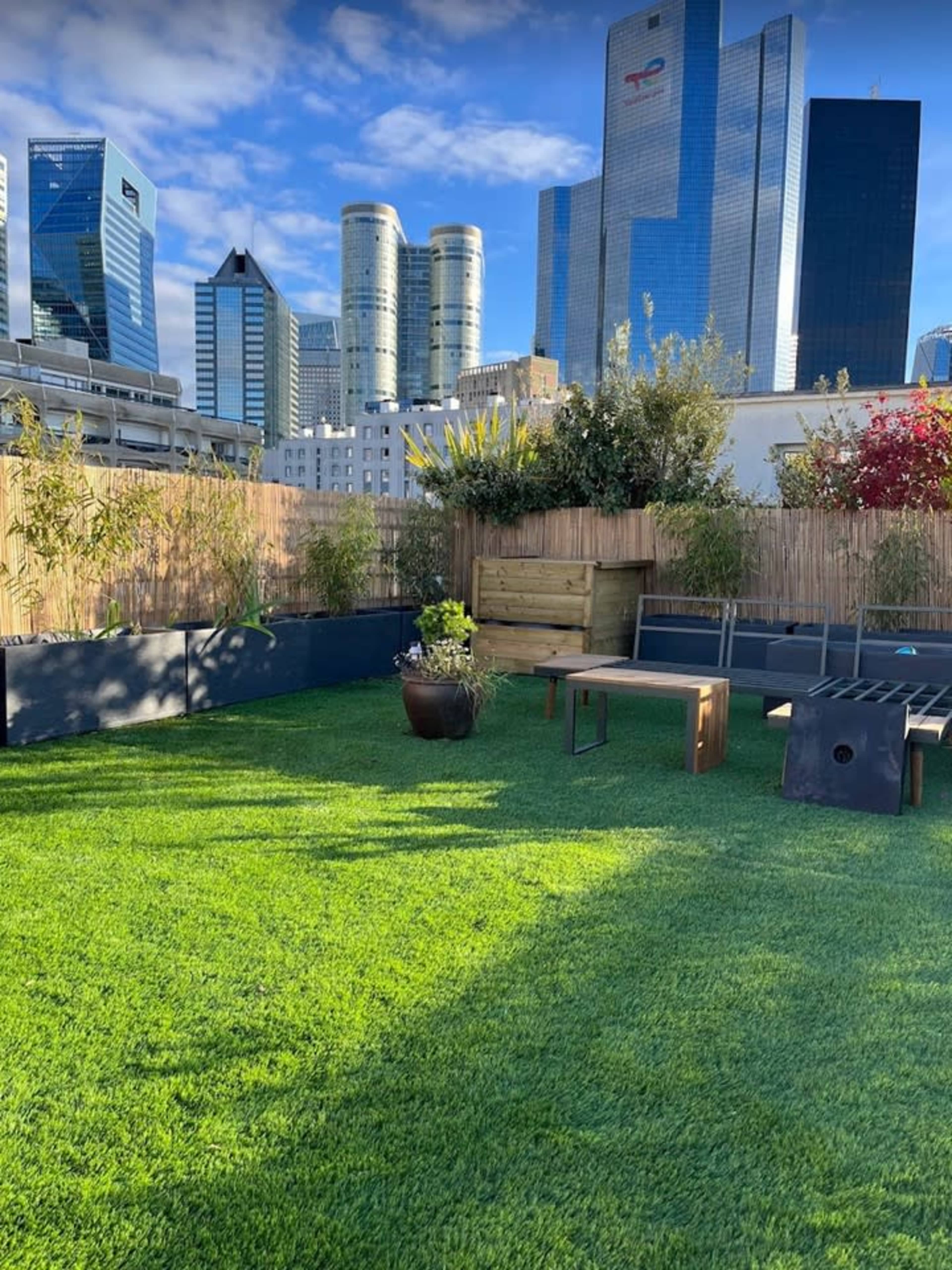 The image shows a rooftop garden with artificial grass, wooden seating, and a backdrop of modern skyscrapers.