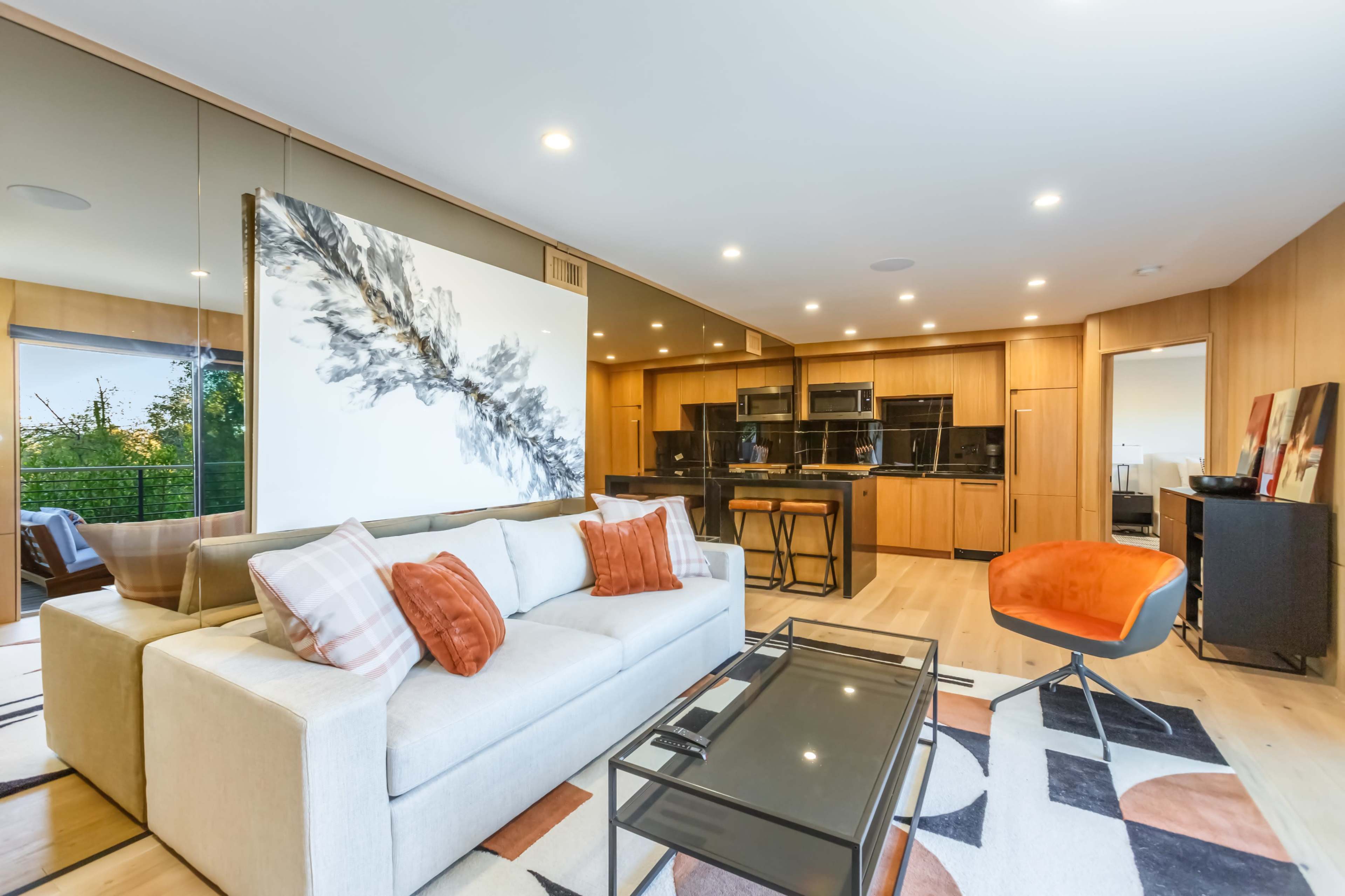 A modern living room featuring a light-colored sofa with orange and white pillows, a glass coffee table, and a stylish mural on the wall, with a dining area visible in the background.