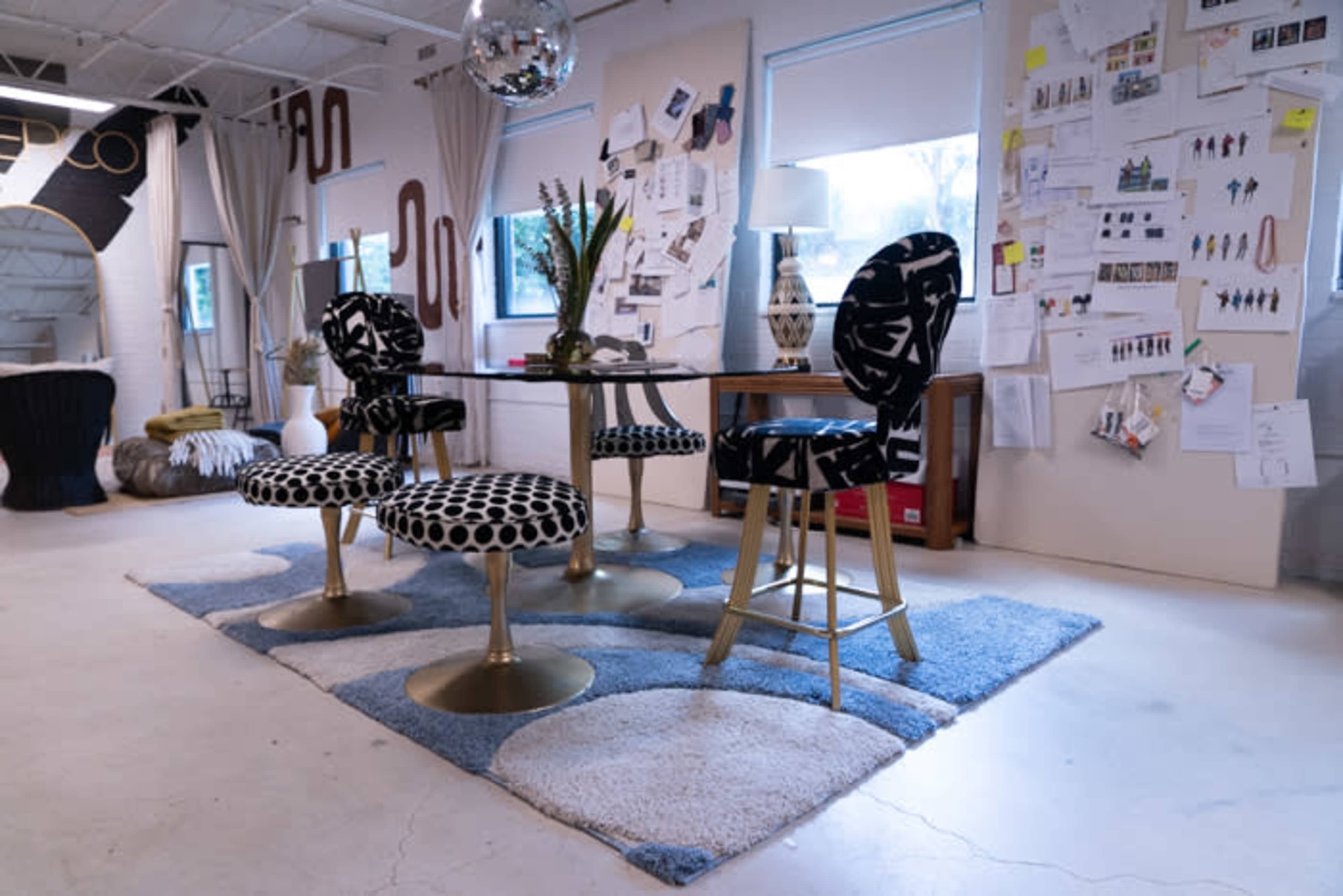 A modern interior design space featuring a round table surrounded by five uniquely patterned chairs on a patterned rug, with design inspiration boards displayed on the walls.