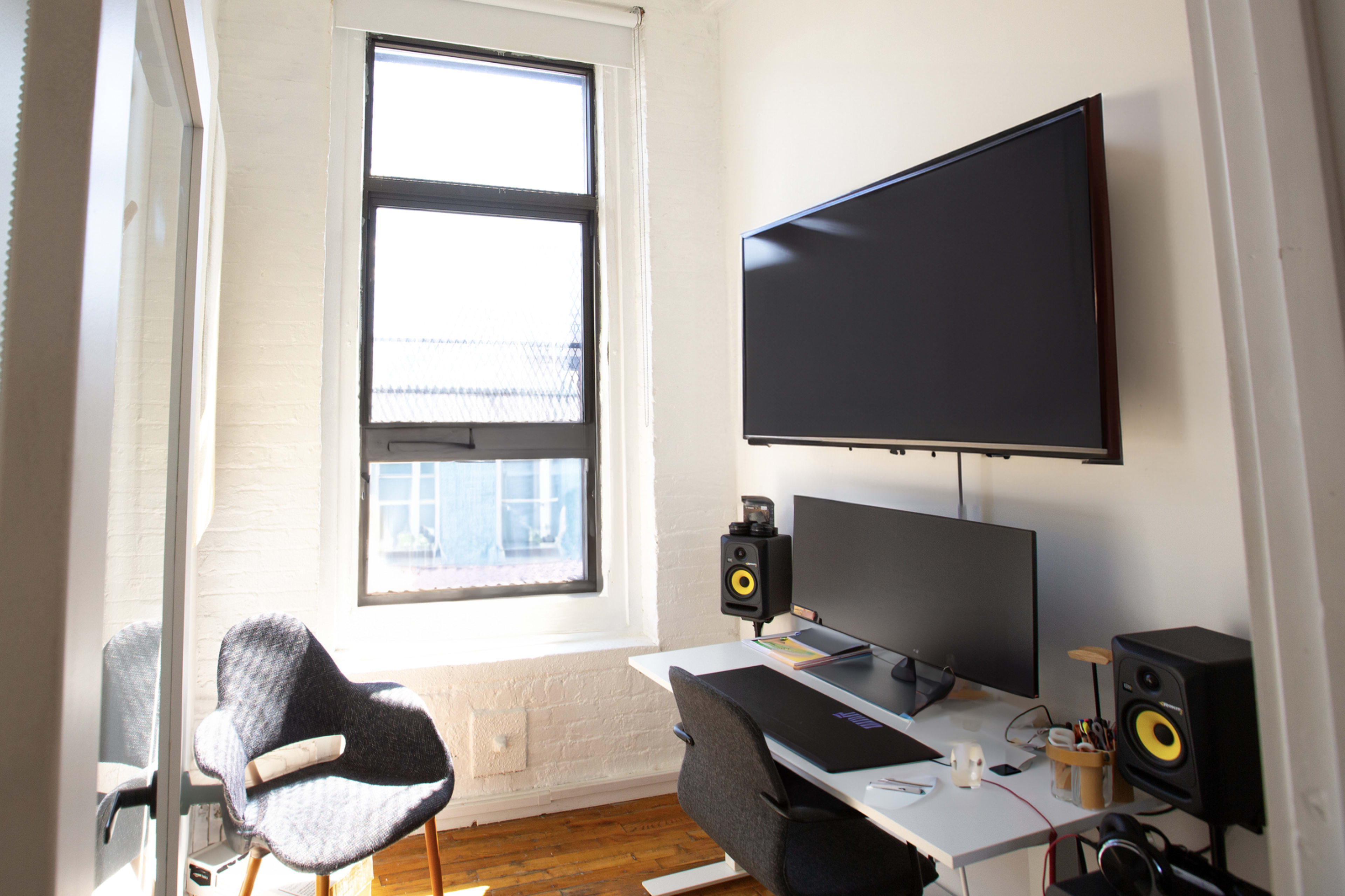 The image shows a bright, minimalist home office with a large wall-mounted television, a desk with dual monitors, and a cozy chair beside a window.