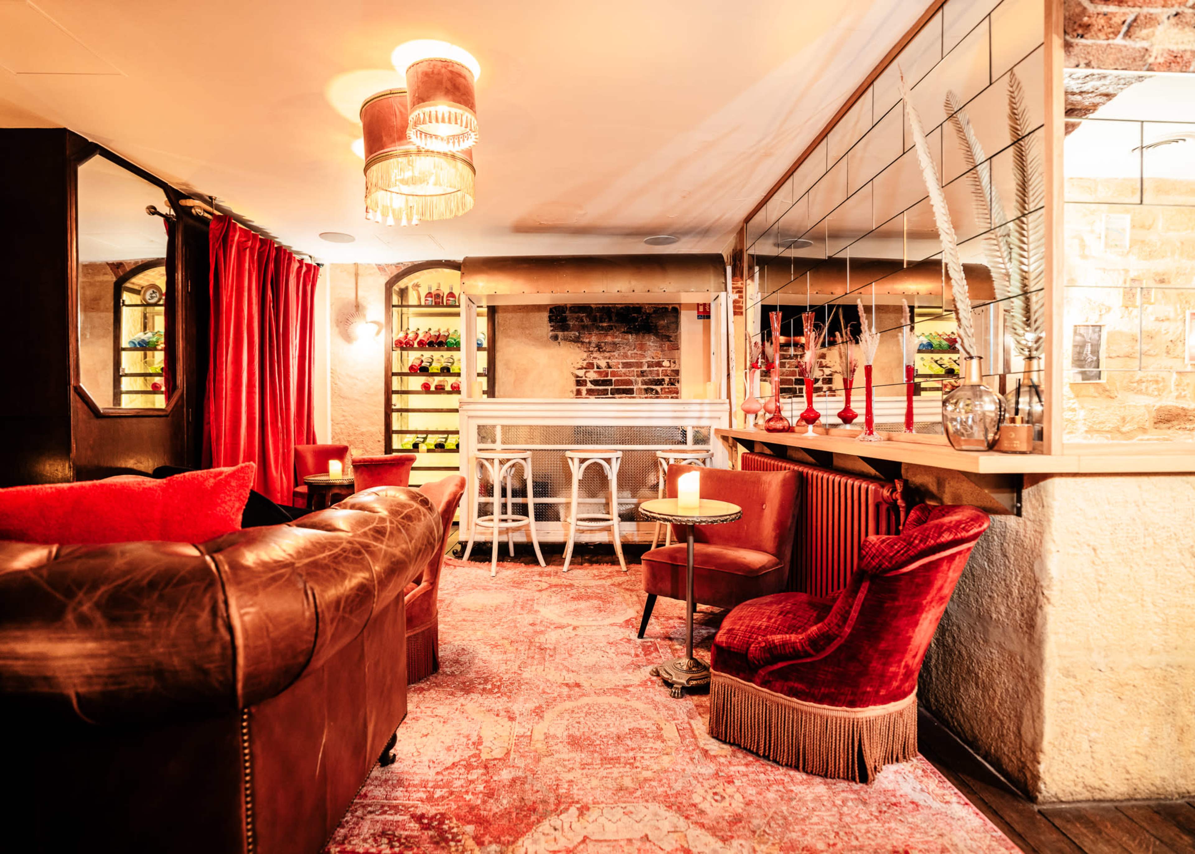 A cozy lounge area featuring a brown leather sofa, a plush red chair, and a small round table, all set against a backdrop of warm lighting and exposed brick walls.