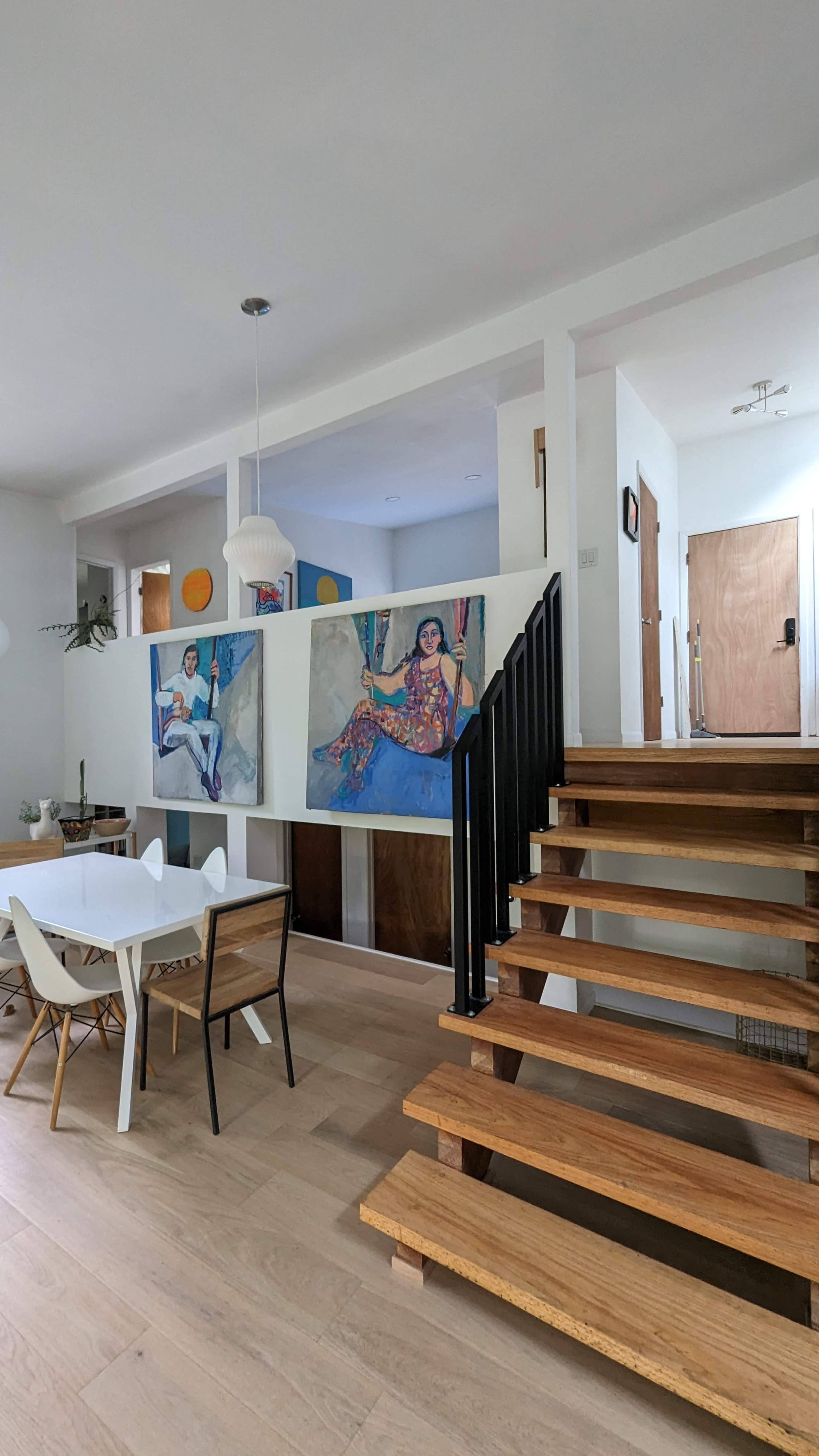 The image shows an interior space featuring a wooden staircase, a dining table with chairs, and colorful artwork displayed on the walls.