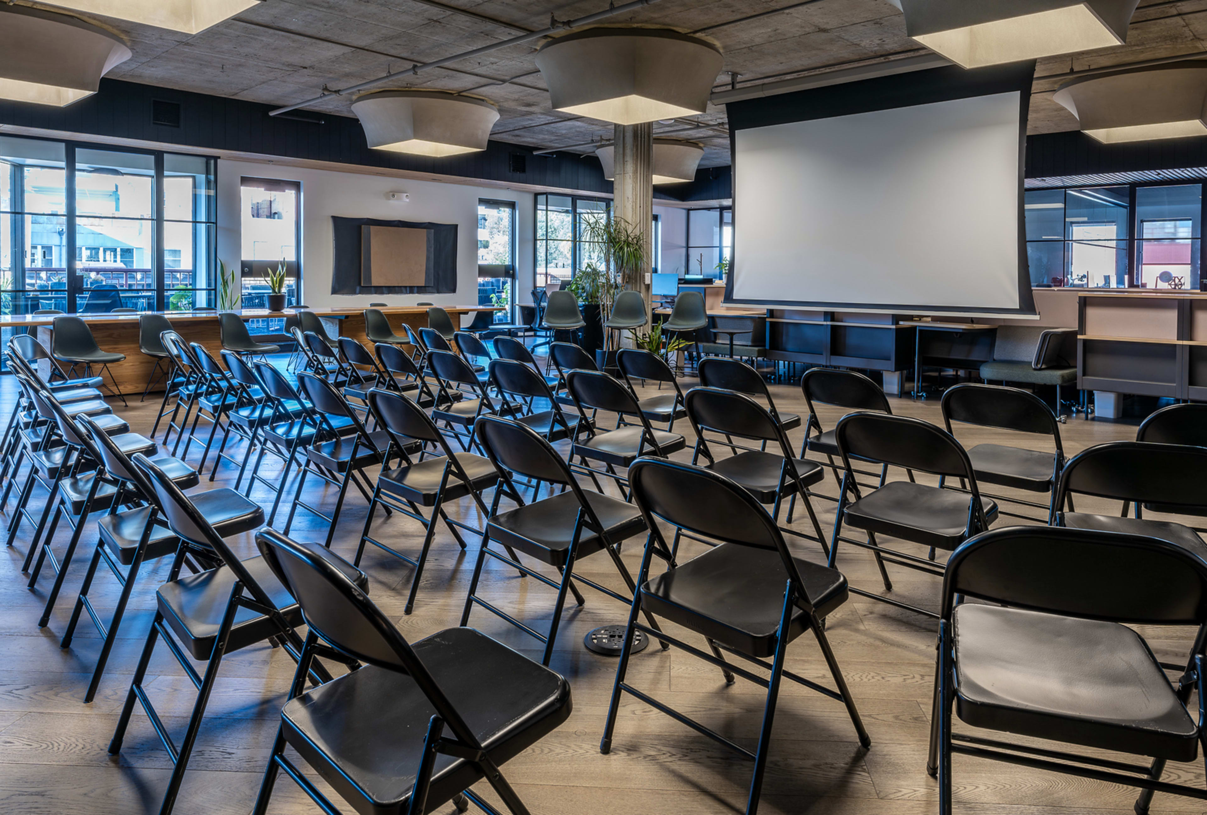 The image shows a modern conference room arranged with rows of black folding chairs facing a large projection screen.