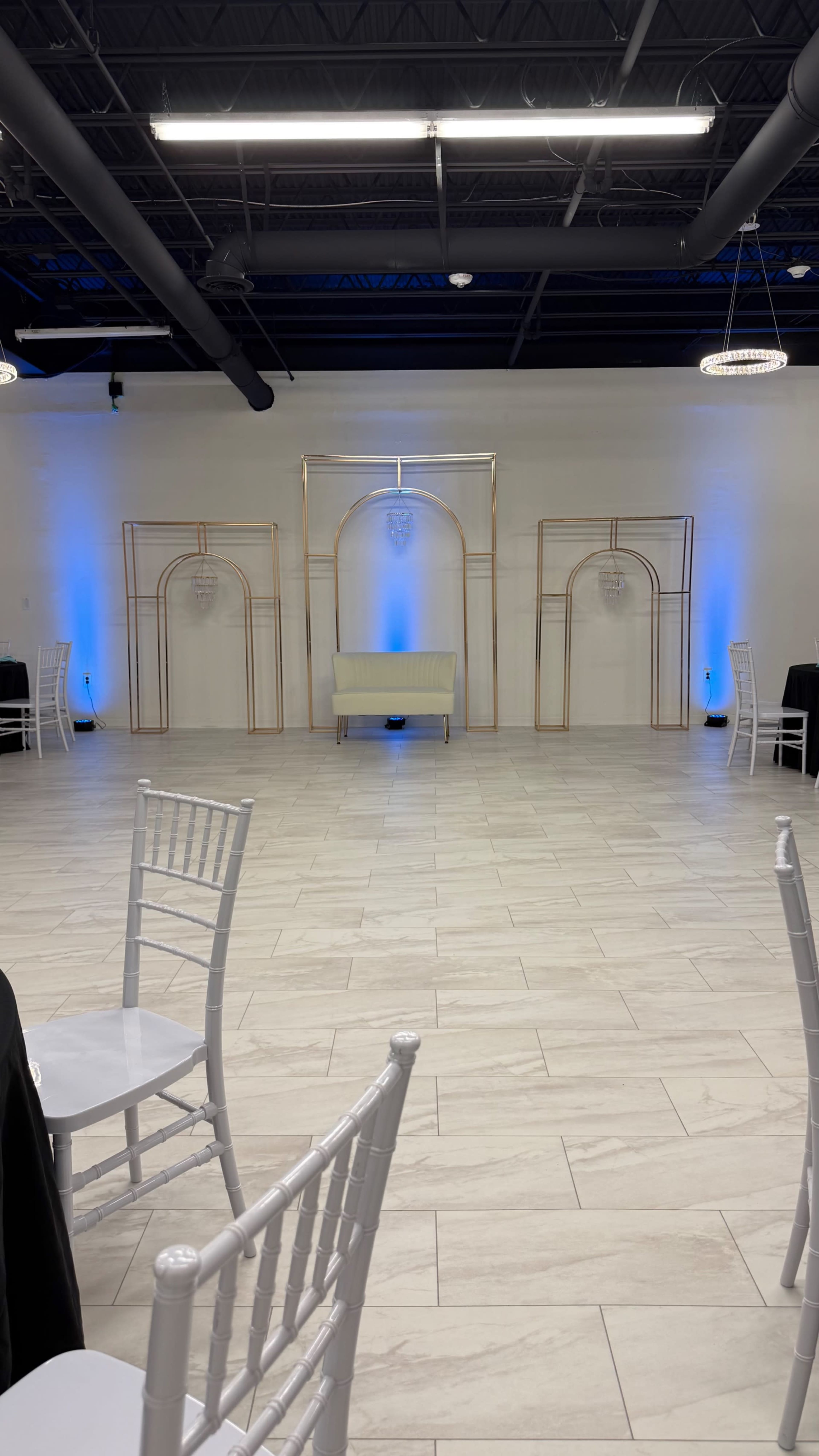 The image shows a spacious event venue with a light-colored floor, featuring a central seating area and decorative arches illuminated by blue lights.