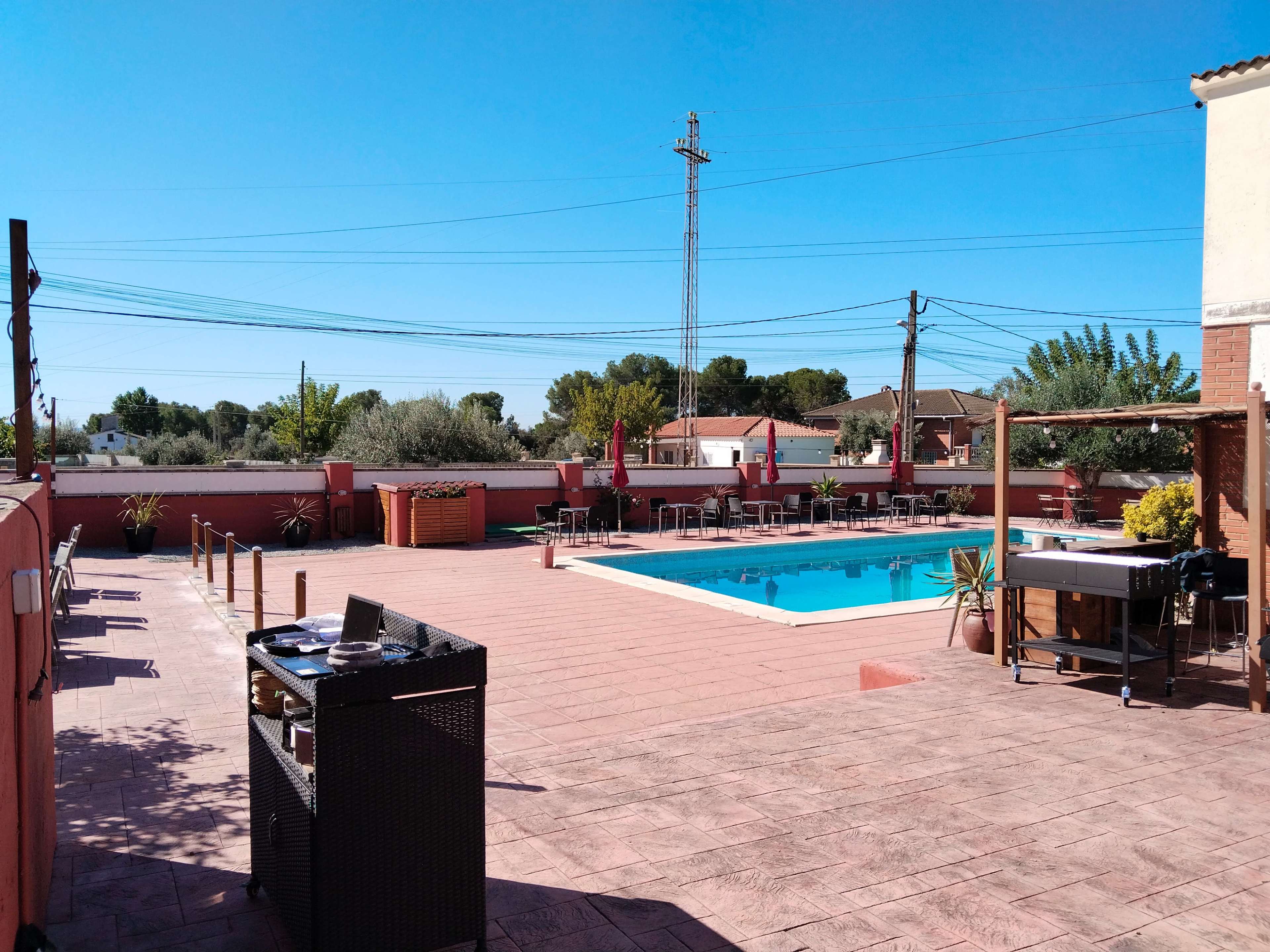 The image shows a spacious outdoor area featuring a swimming pool surrounded by lounge chairs, tables, and greenery under a clear blue sky.
