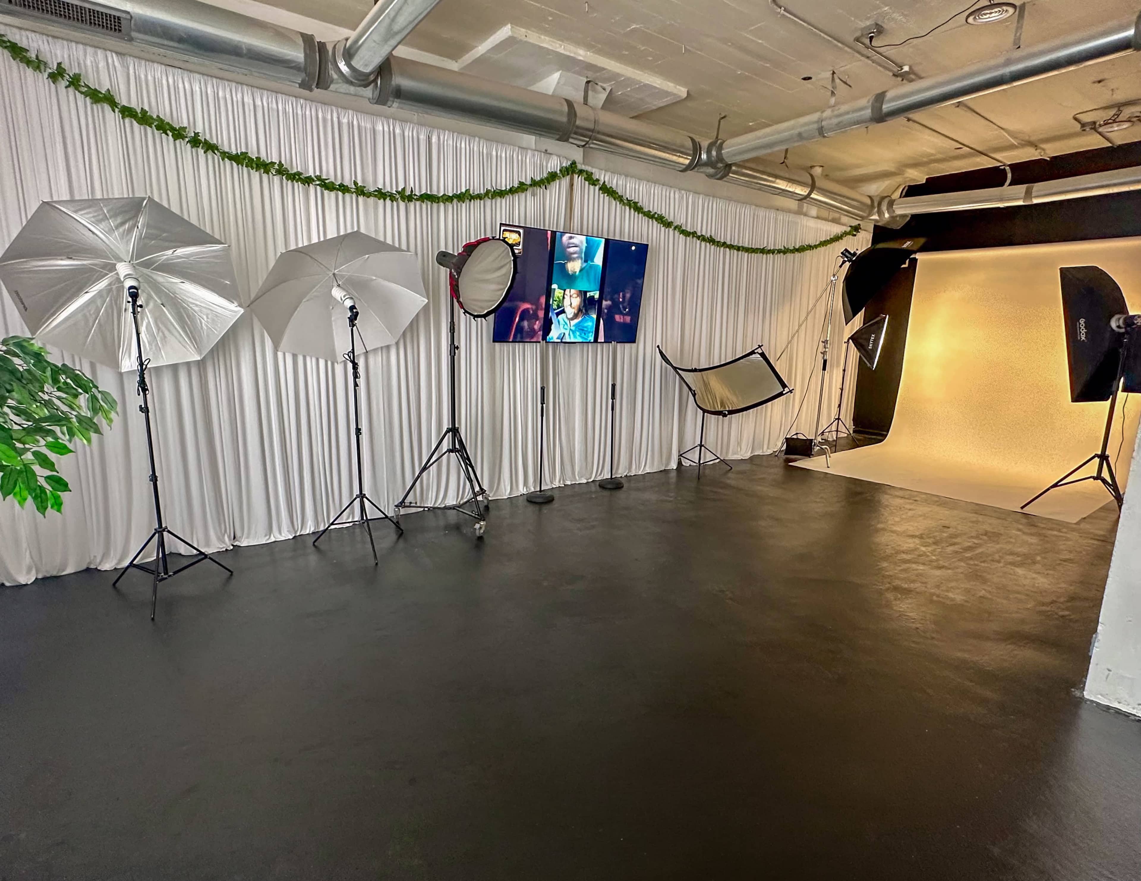 The image shows a photography studio with multiple lighting equipment, a large display screen, and a plain background setup against an abstract wall.