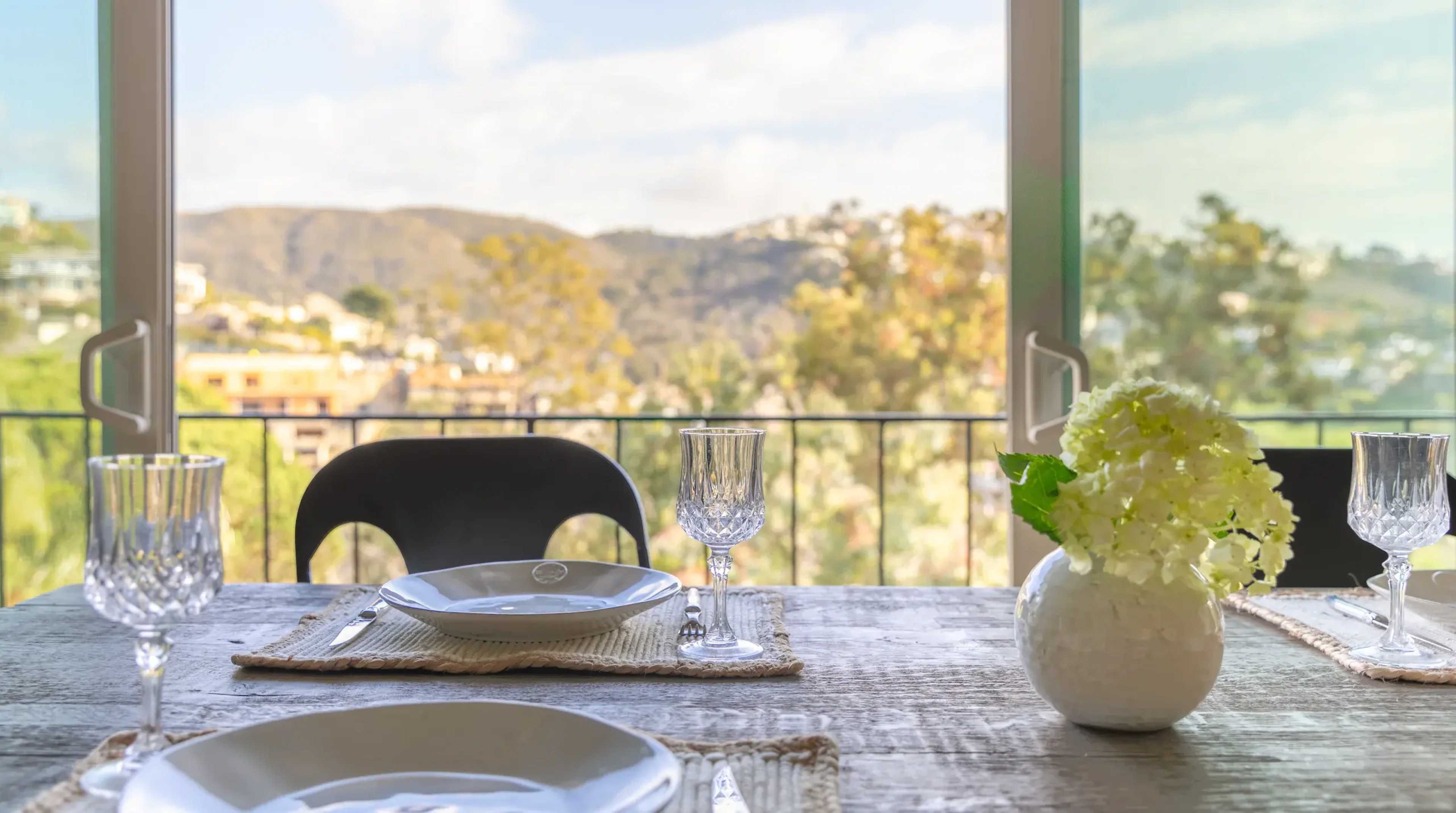 A dining table is set with plates and glassware, overlooking a scenic view of hills and trees through large windows.