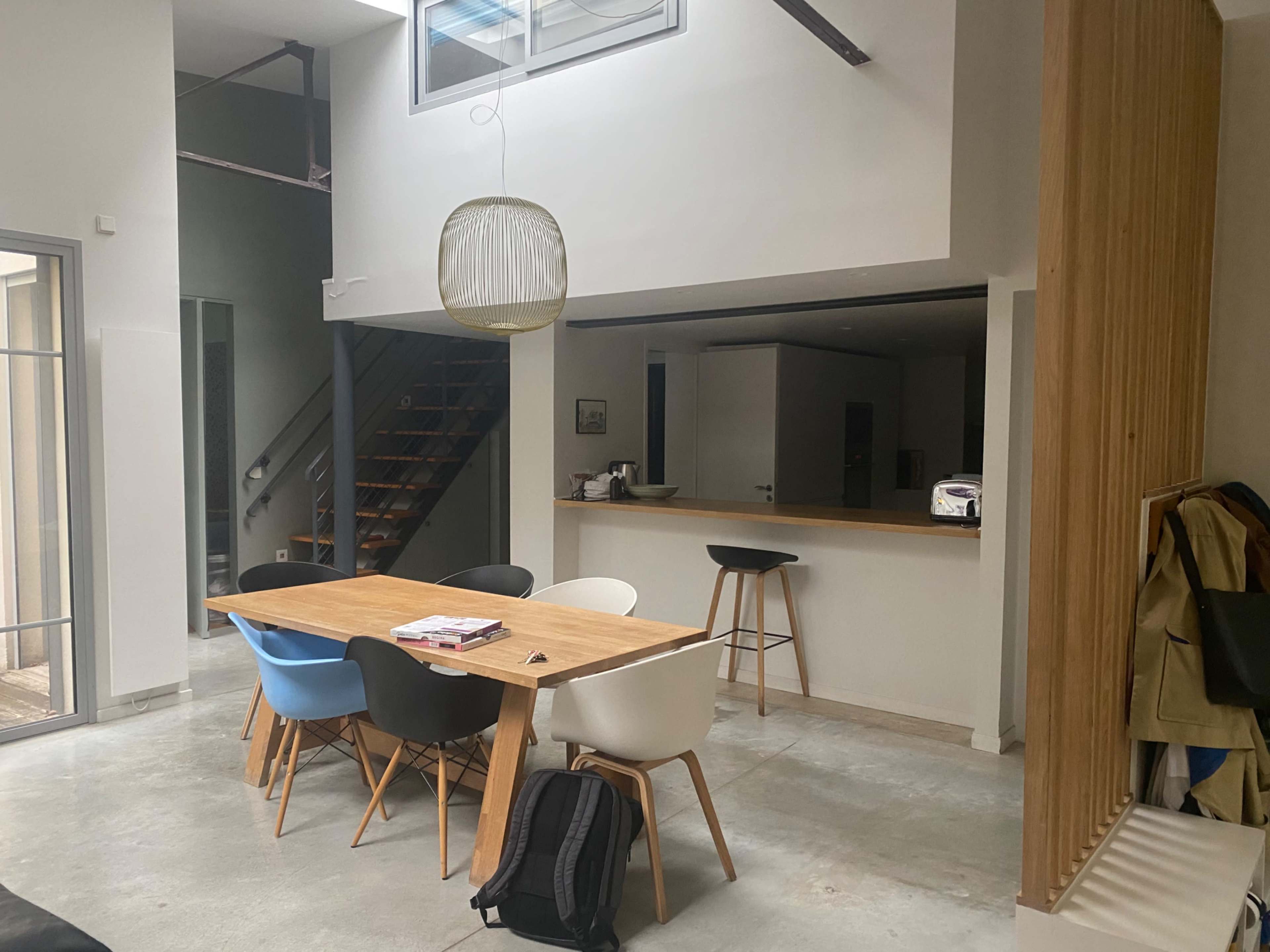 A modern, open-concept dining area with a wooden table surrounded by mismatched chairs, leading to a staircase and kitchen area.