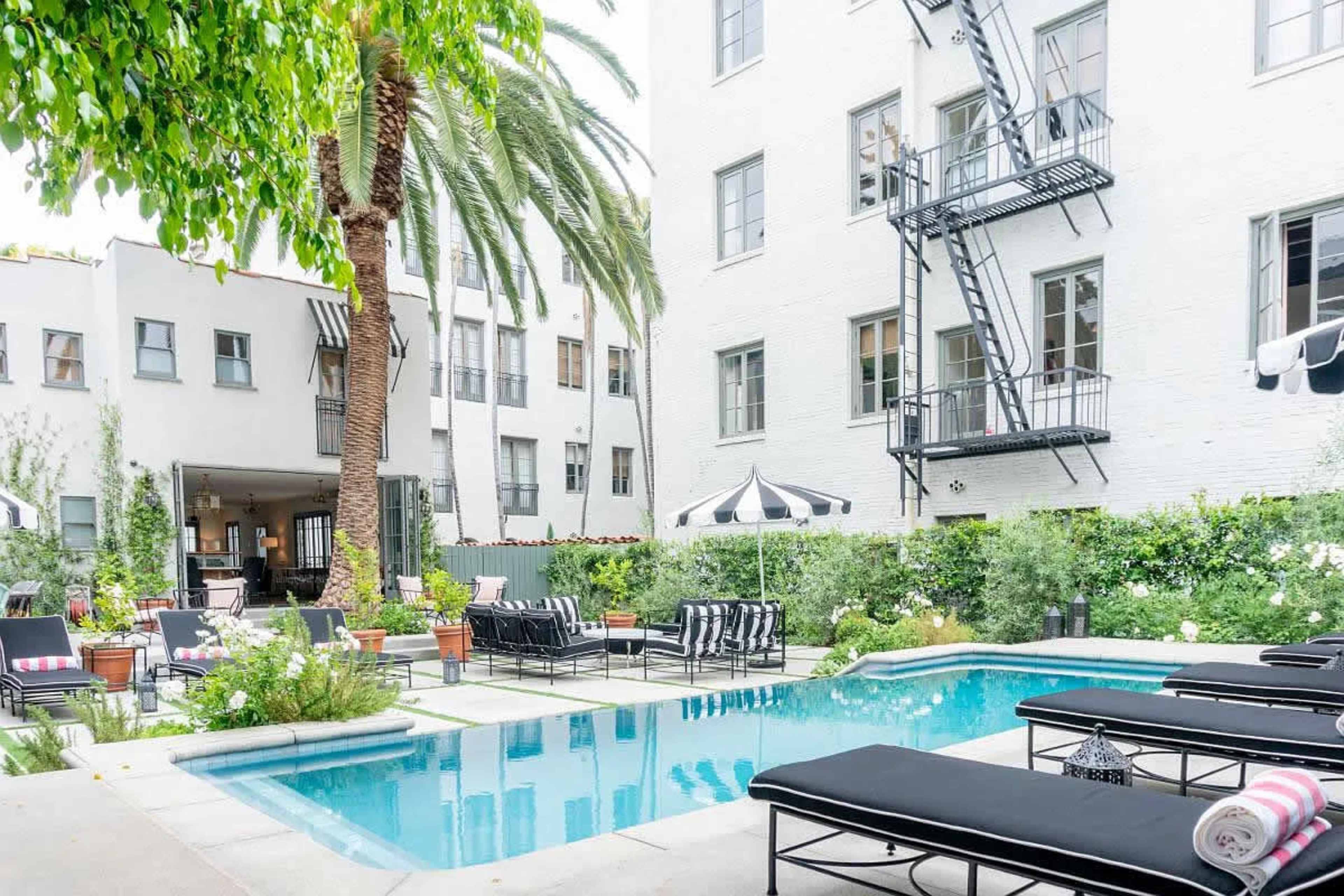 The image shows a swimming pool surrounded by lounge chairs, palm trees, and greenery in a courtyard of a multi-story building.