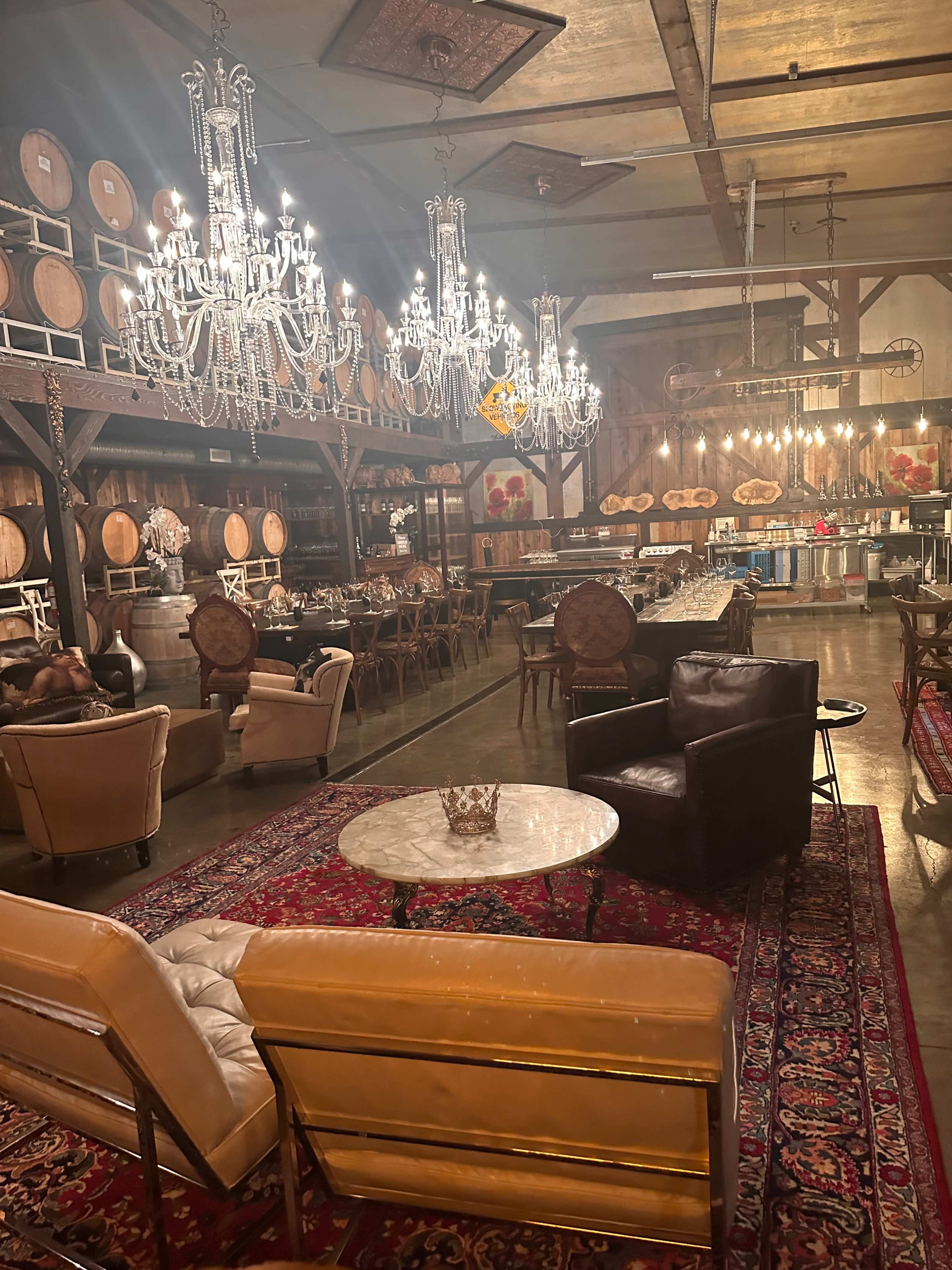 The image shows an elegant rustic event space featuring chandeliers, wine barrels, and a mix of comfortable seating arrangements.