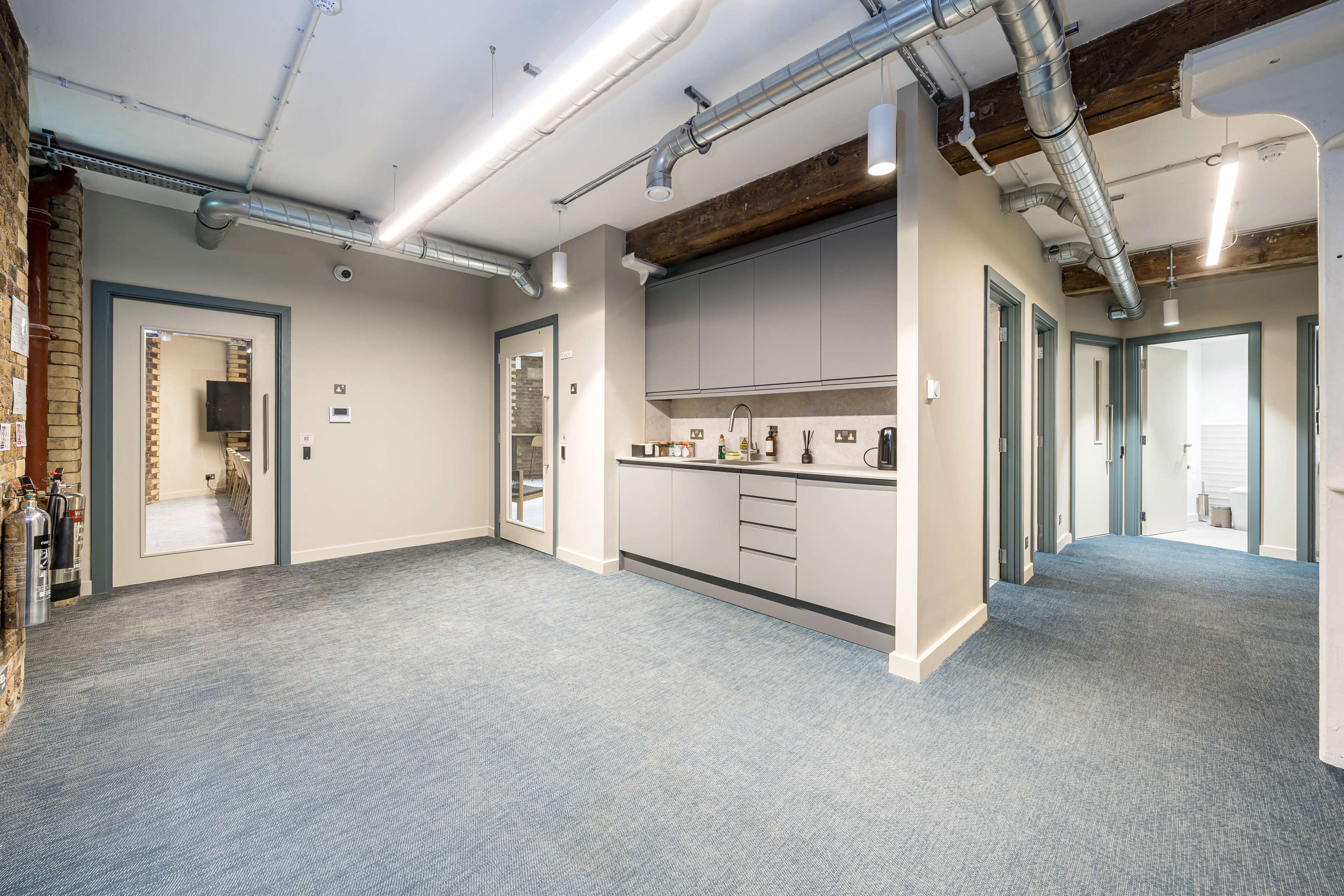 The image shows a modern kitchen area with gray cabinets and a blue carpeted floor, adjacent to two doorways leading to separate rooms.