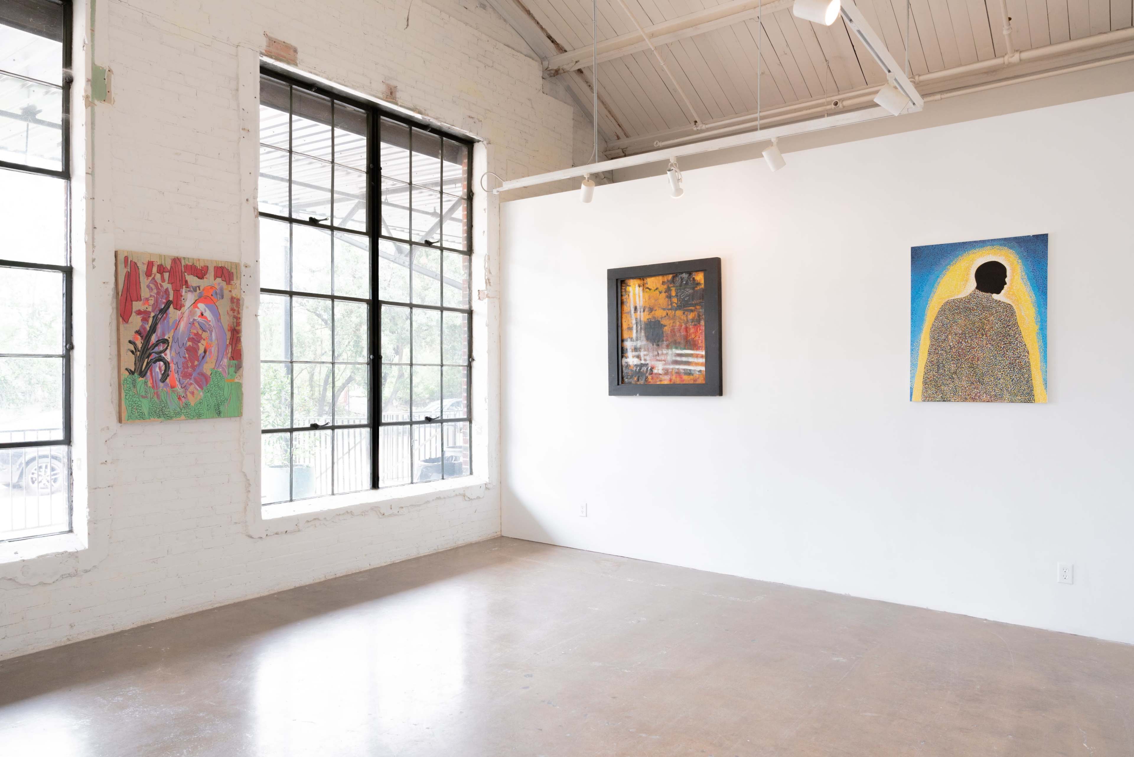 The image shows an interior art gallery with three paintings displayed on a white wall and large windows letting in natural light.