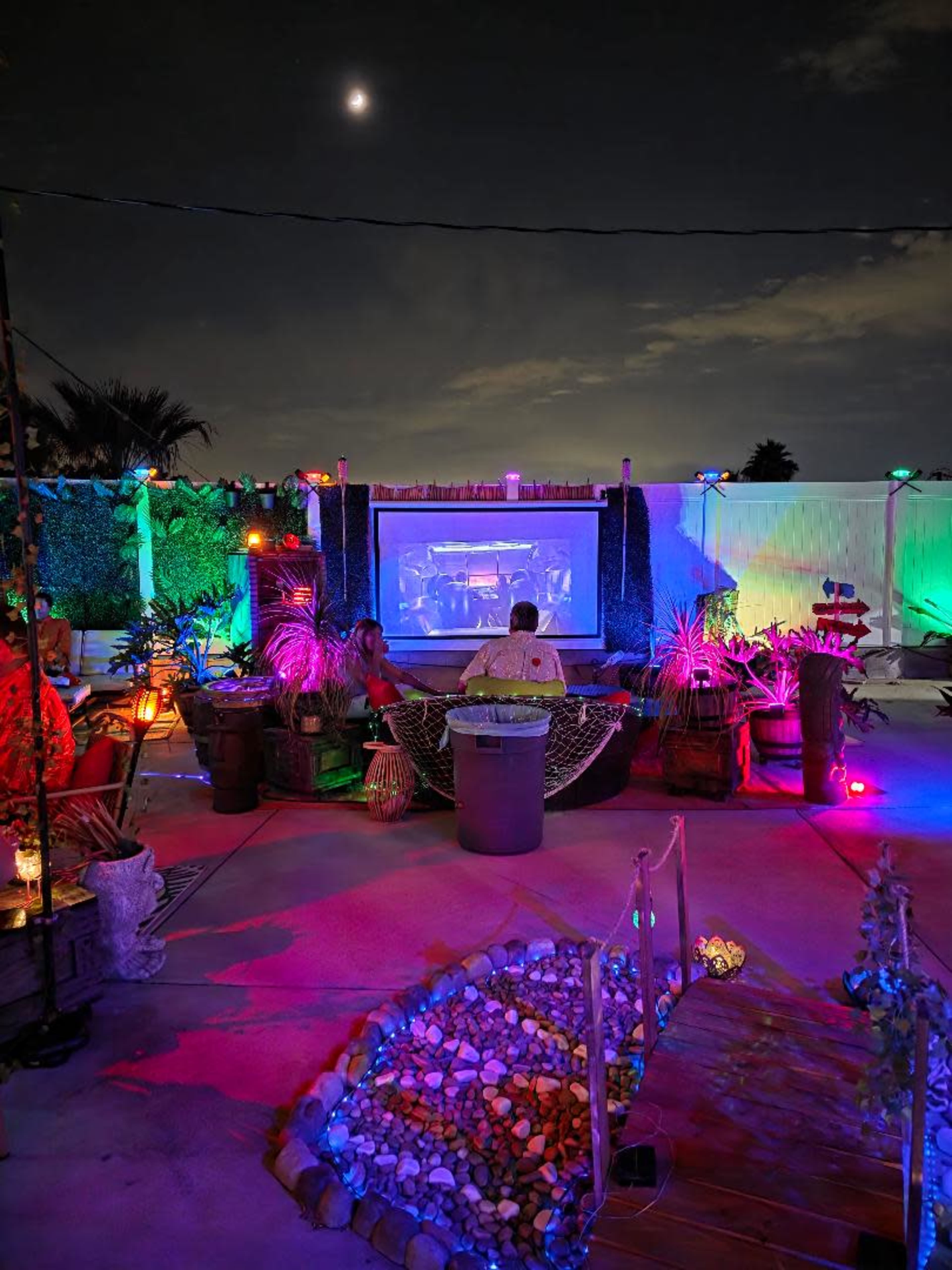 A brightly lit outdoor space features a movie screen surrounded by plants, decorative lighting, and seating areas under a twilight sky.