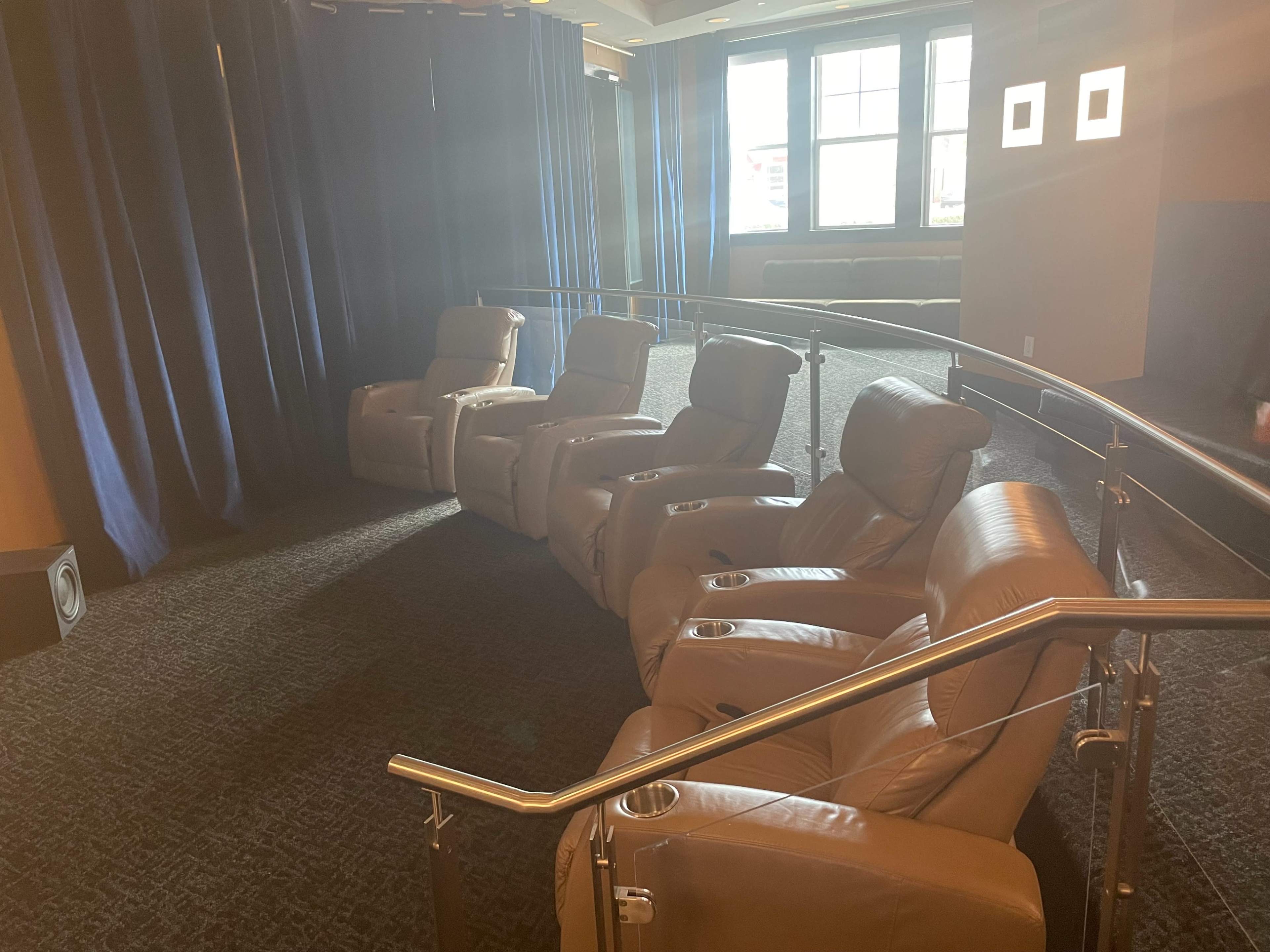 A row of beige reclining seats with cup holders, arranged on a carpeted floor in a room with blue curtains and windows in the background.