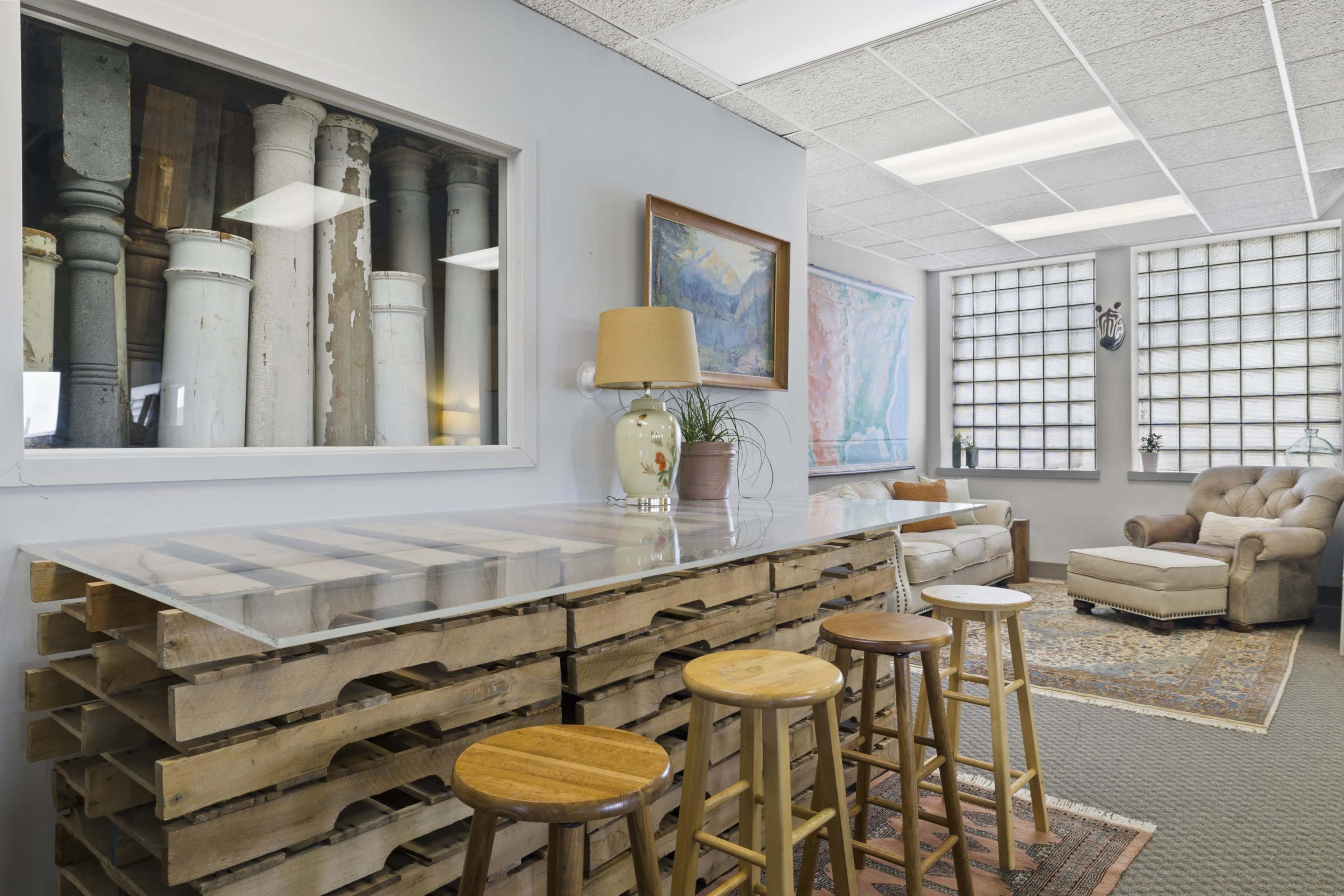 The scene shows a modern interior space featuring a wooden pallet bar with four stools, a cozy armchair, and large windows with natural light.