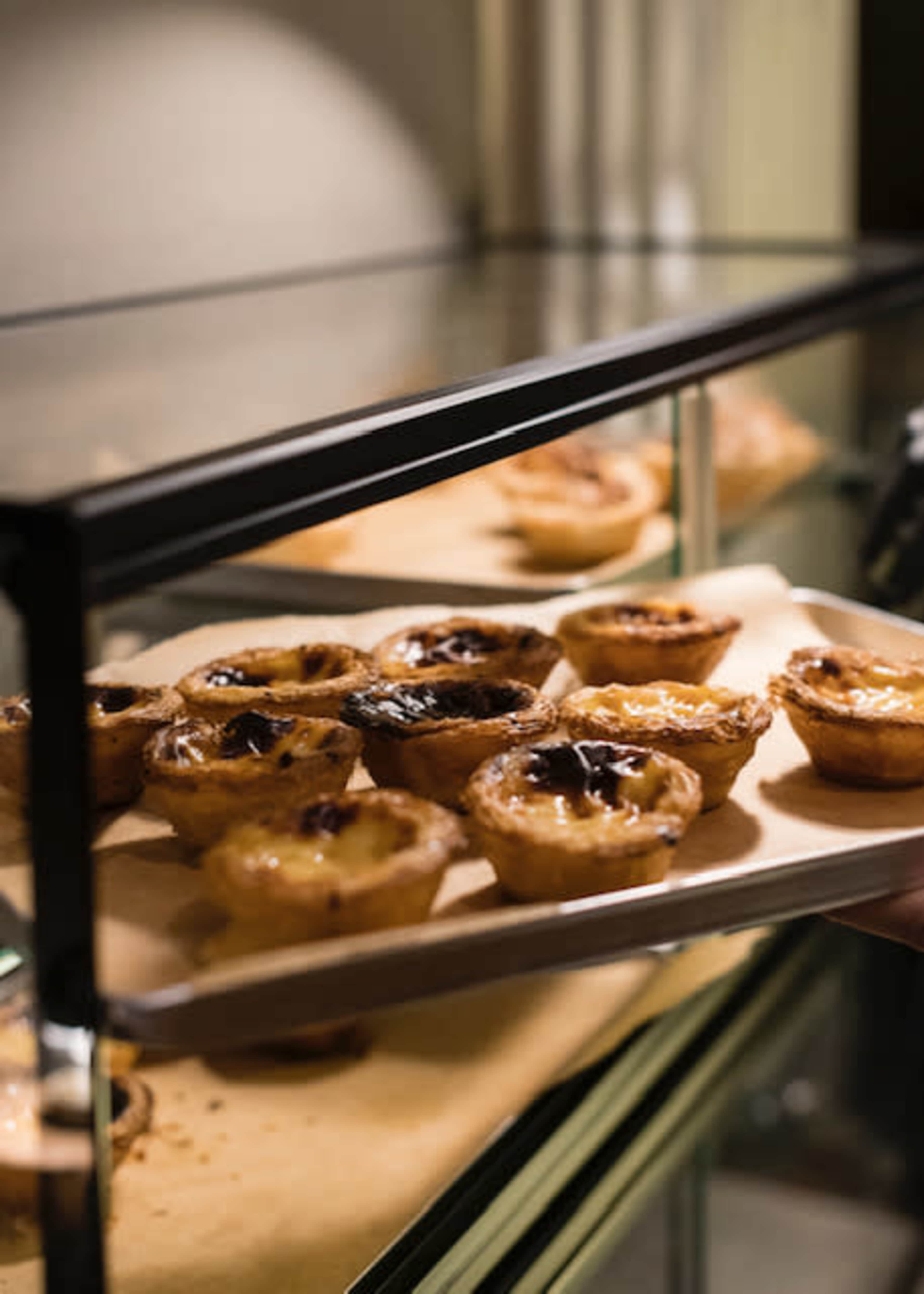 A tray of baked pastries sits on a glass display shelf, showcasing an assortment of tarts with golden-brown crusts.